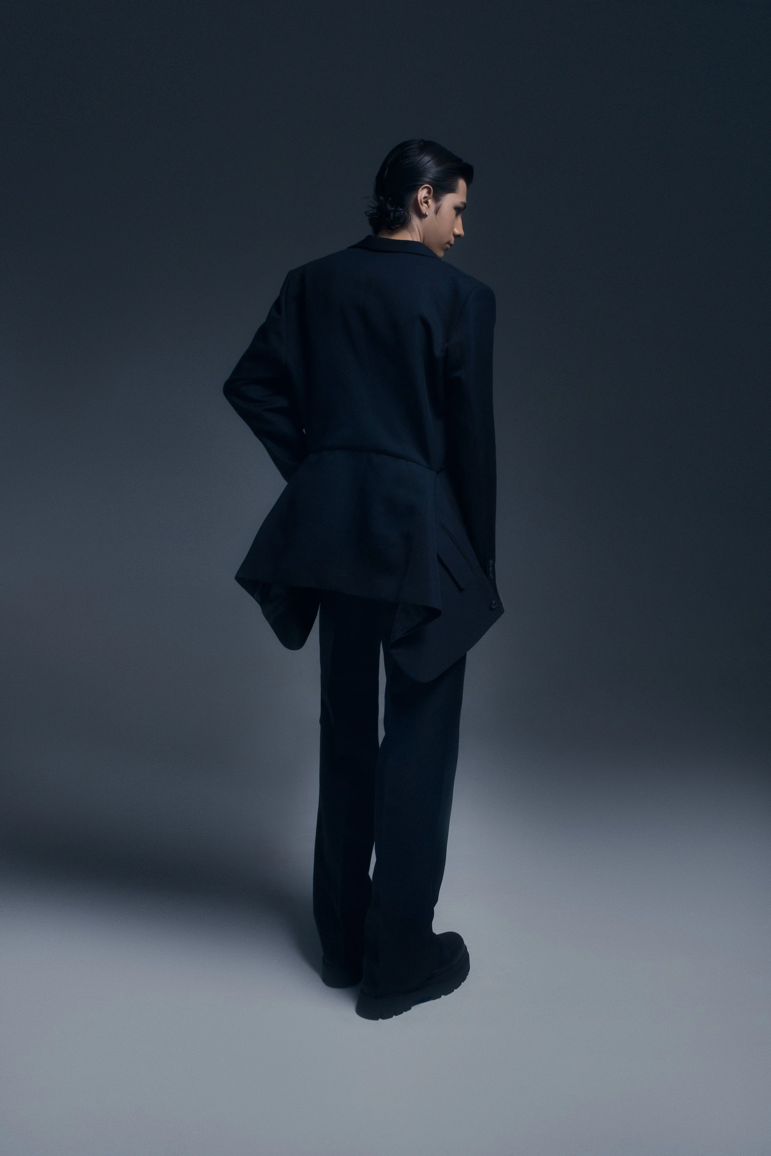 Persona vestida con traje negro, de espaldas, en un fondo gris oscuro.