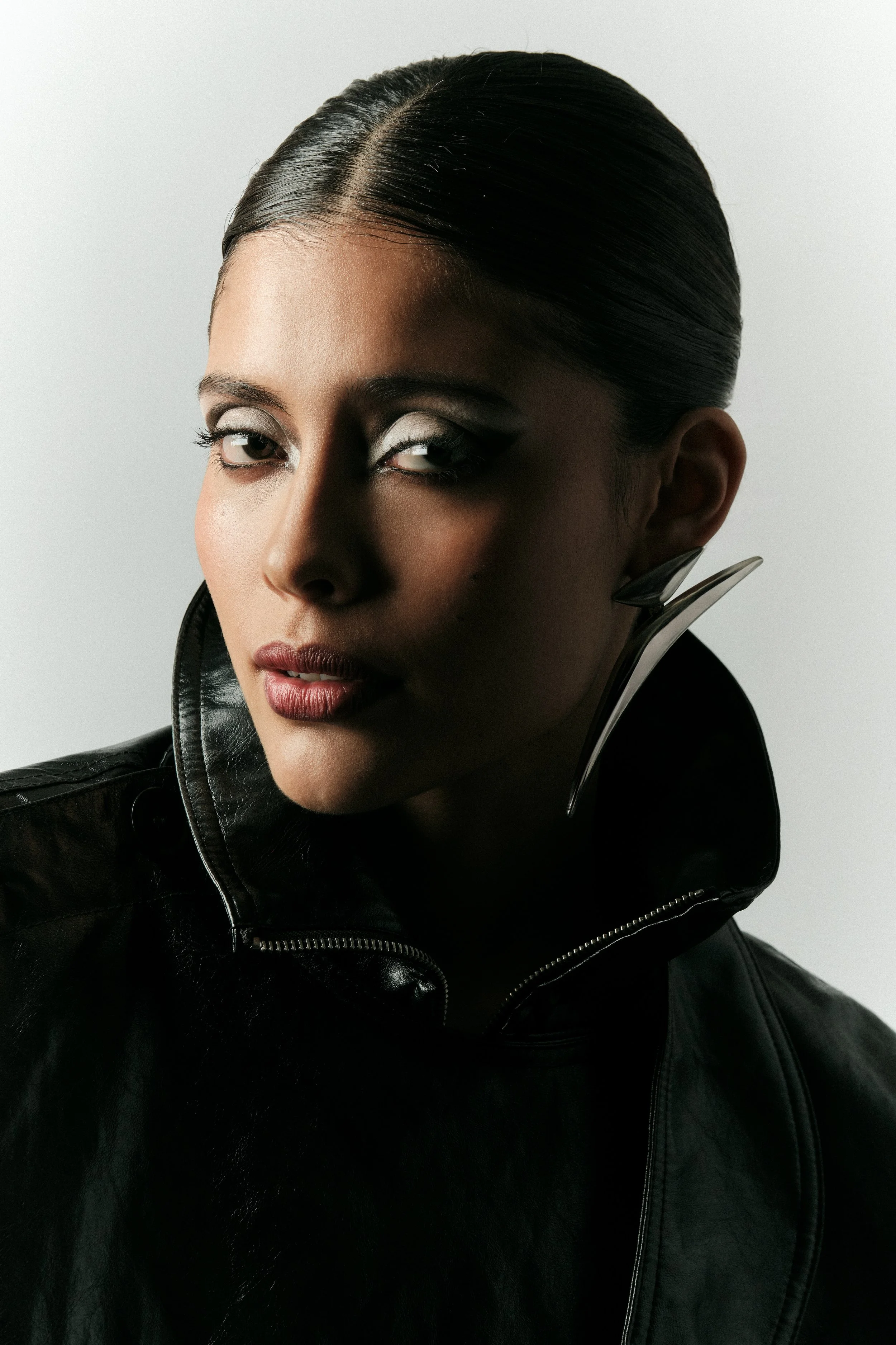 Mujer con cabello liso y negro, usa maquillaje con sombra plateada, veste una chaqueta de cuero negra con el cuello levantado y un pendiente grande en forma de cuchara.