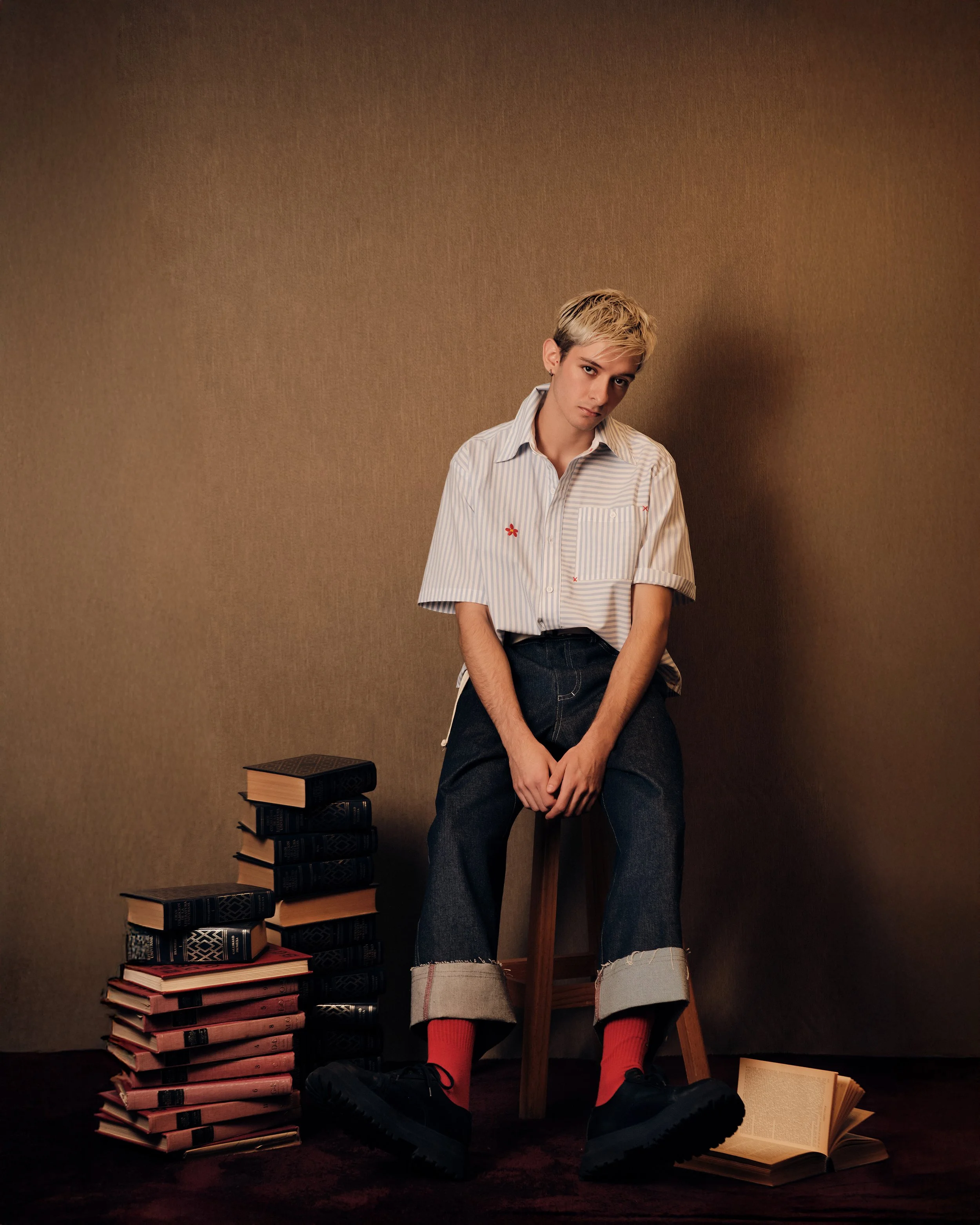 Joven sentado en una banca de madera en una habitación con pared marrón, rodeado de pilas de libros y unos sobre el suelo, con pantalones cortos, camiseta a rayas, calcetas rojas y zapatos negros.