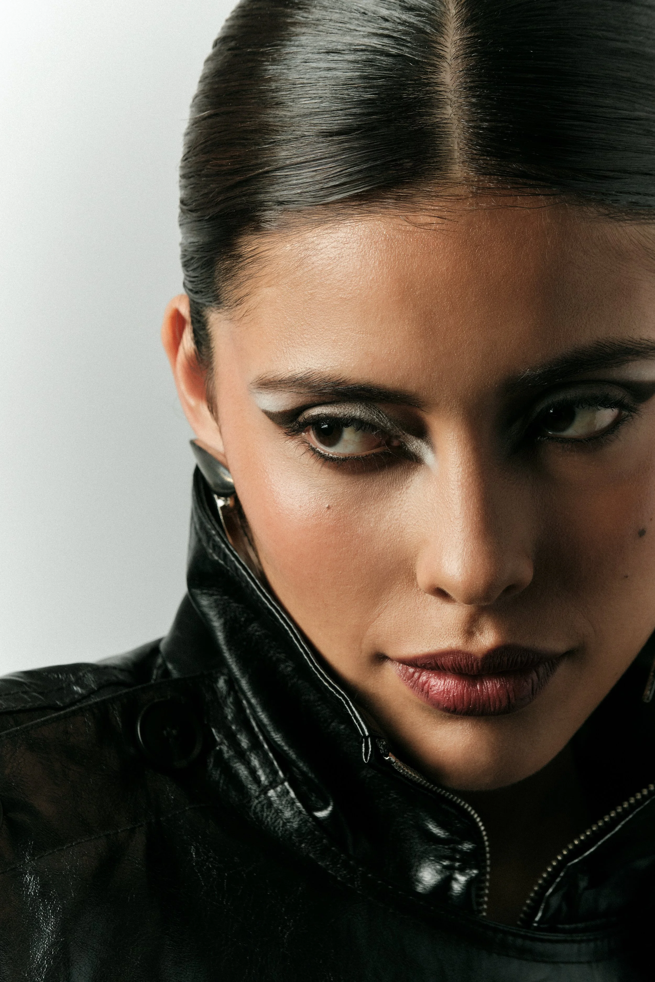 Primer plano de una mujer con maquillaje de ojos oscuro y labios rojos, con cabello oscuro peinado hacia atrás y usando chaqueta de cuero negro.