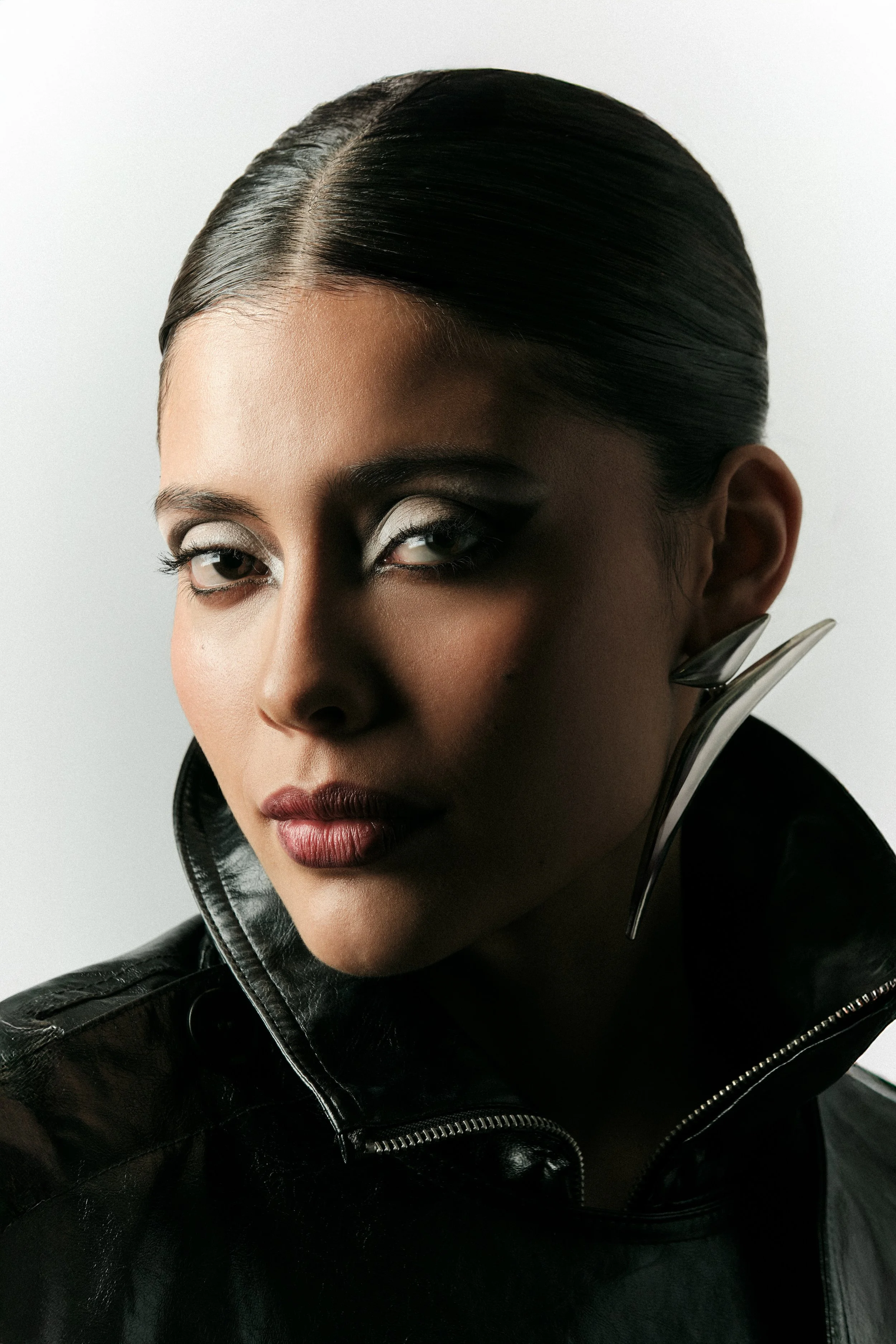 Retrato de una mujer con cabello liso y oscuro, maquillaje llamativo, ropa de cuero negro y un pendiente metálico grande.