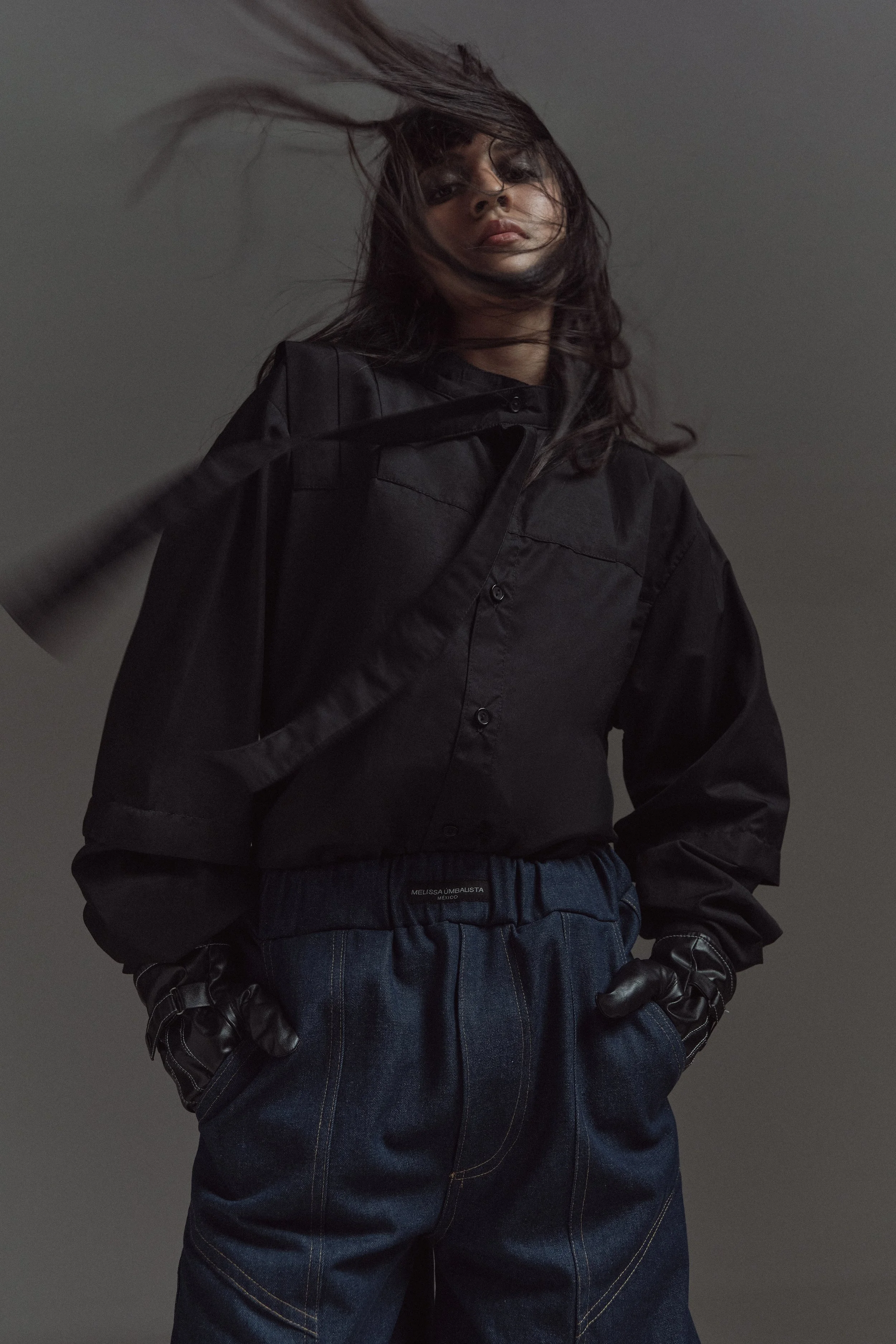 Retrato de una mujer con cabello suelto en movimiento, usando un abrigo negro y guantes de cuero, de origen desconocido.