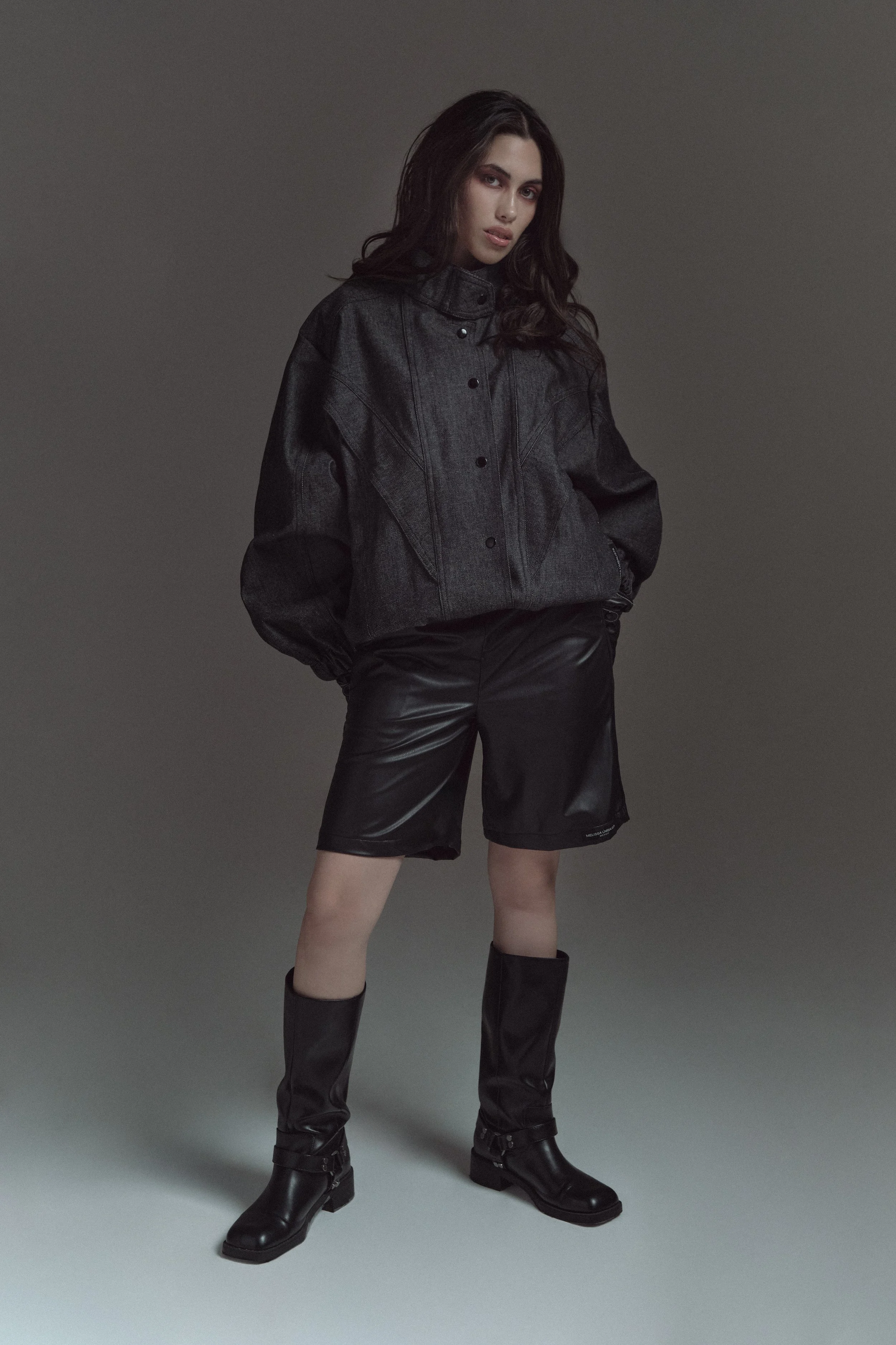 Mujer joven con cabello largo, usando una chaqueta de mezclilla negra, pantalones cortos de cuero y botas negras altas, posando en un fondo gris.