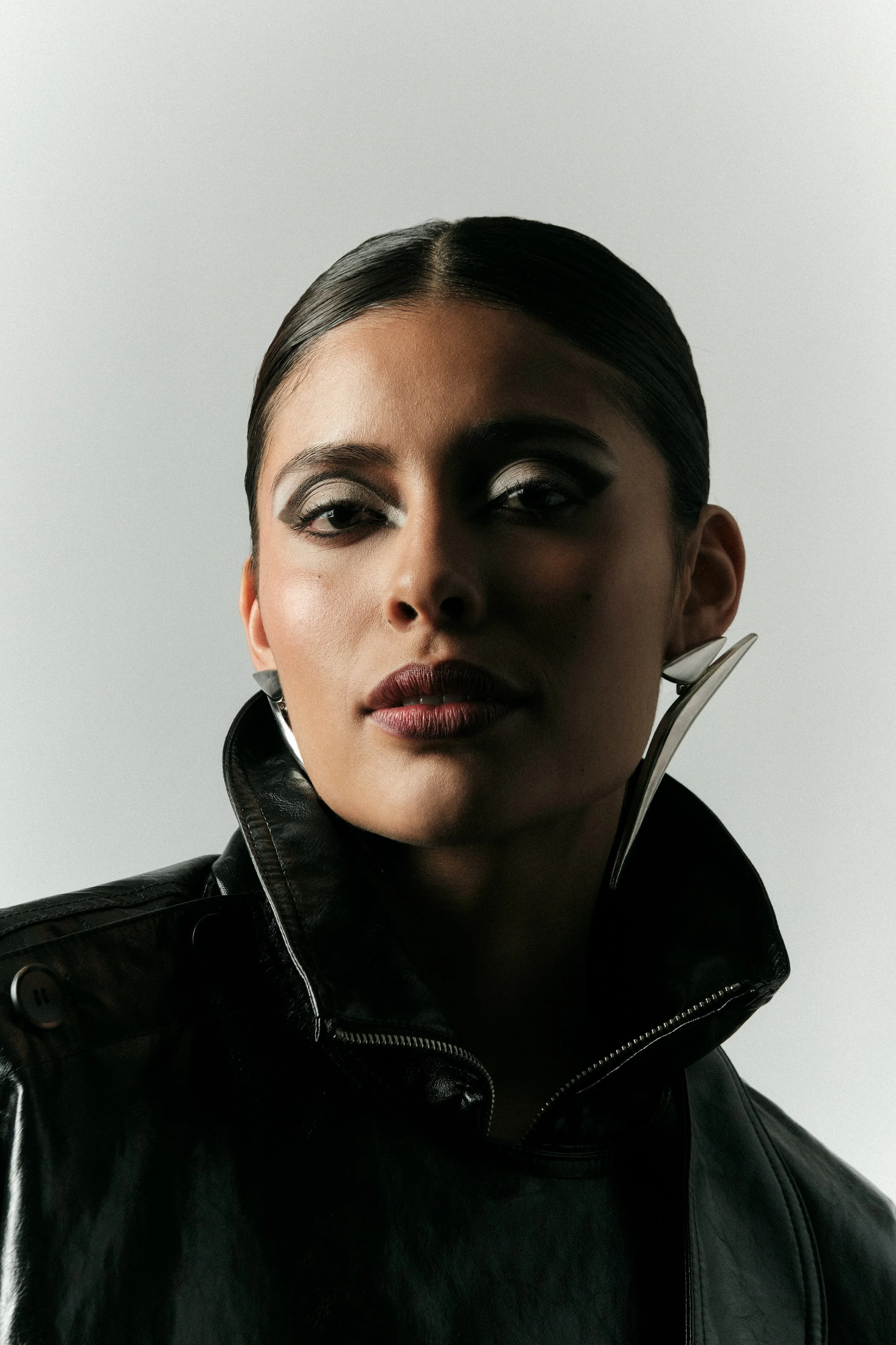 Retrato de una mujer con maquillaje oscuro, cabello peinado hacia atrás, usa una chaqueta de cuero negra y un pendiente grande de metal, con fondo gris claro.