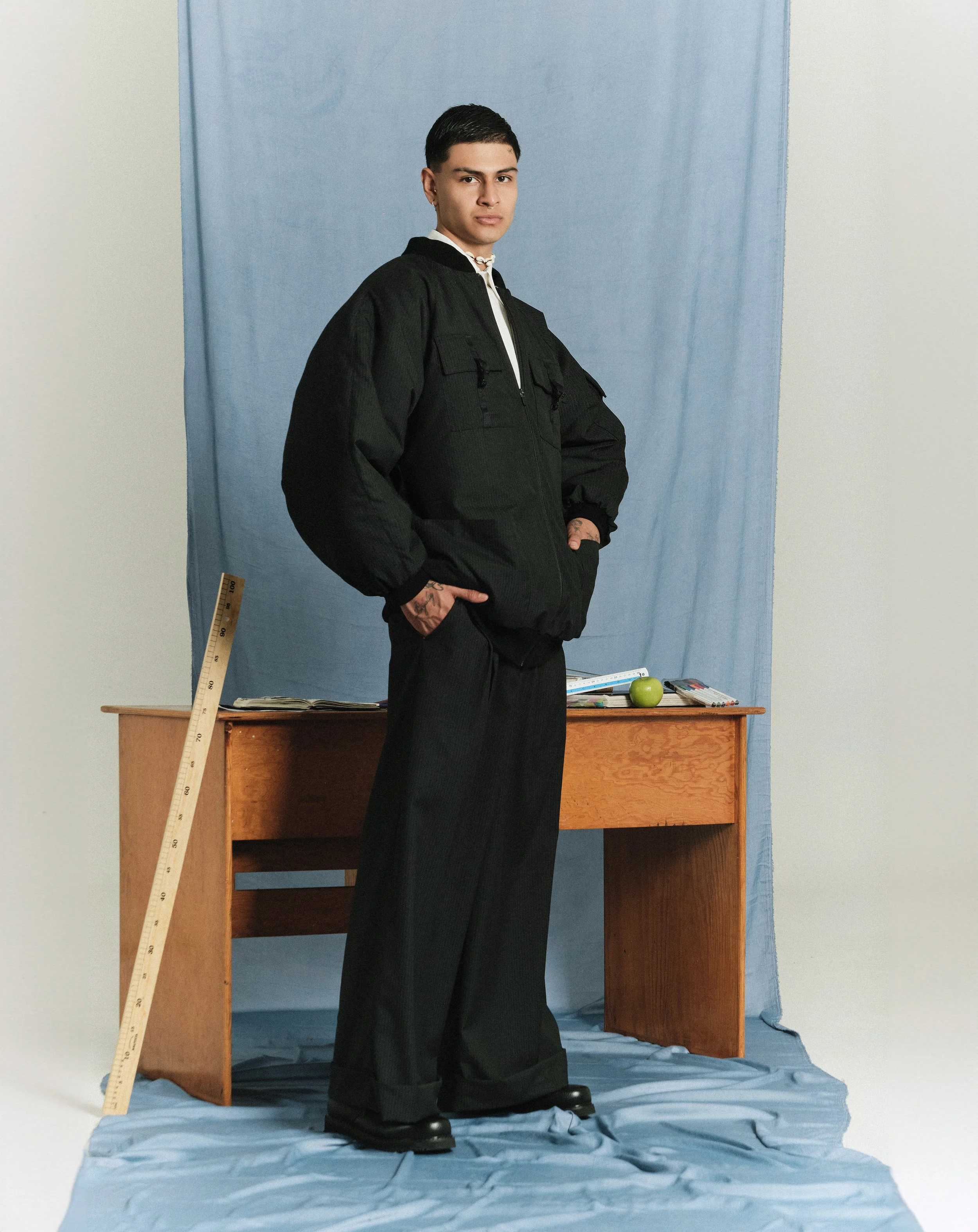 Joven de pie con ropa negra frente a un fondo azul, junto a un escritorio de madera con libros, un pizarrón, una manzana y algunos objetos. Rodeado de tela azul en el suelo y fondo.