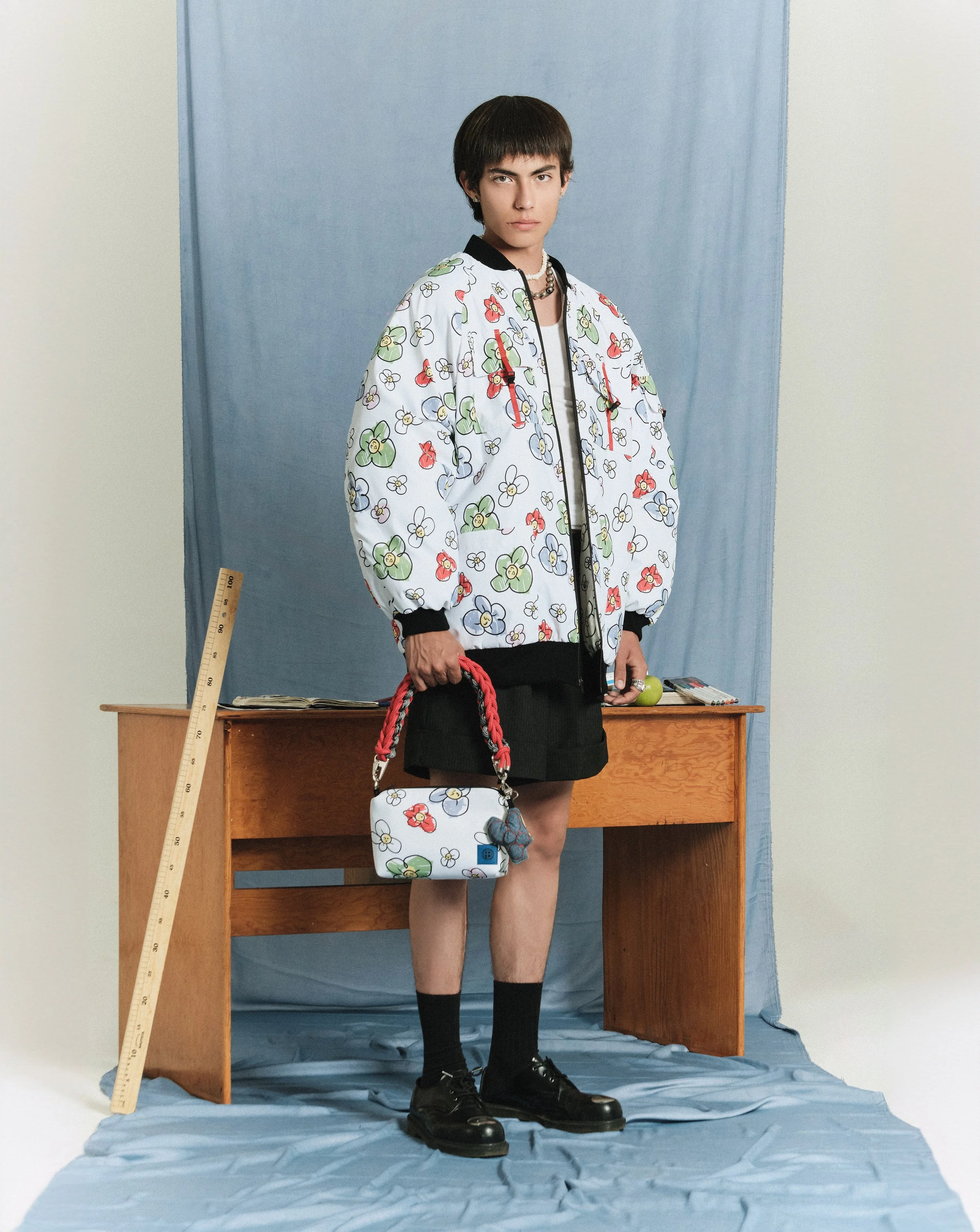 Joven con estilo fashion, lleva una chaqueta con estampado floral, falda negra, mochila a juego, joyería y calcetas negras, con fondo azul y objetos de oficina en una mesa de madera.