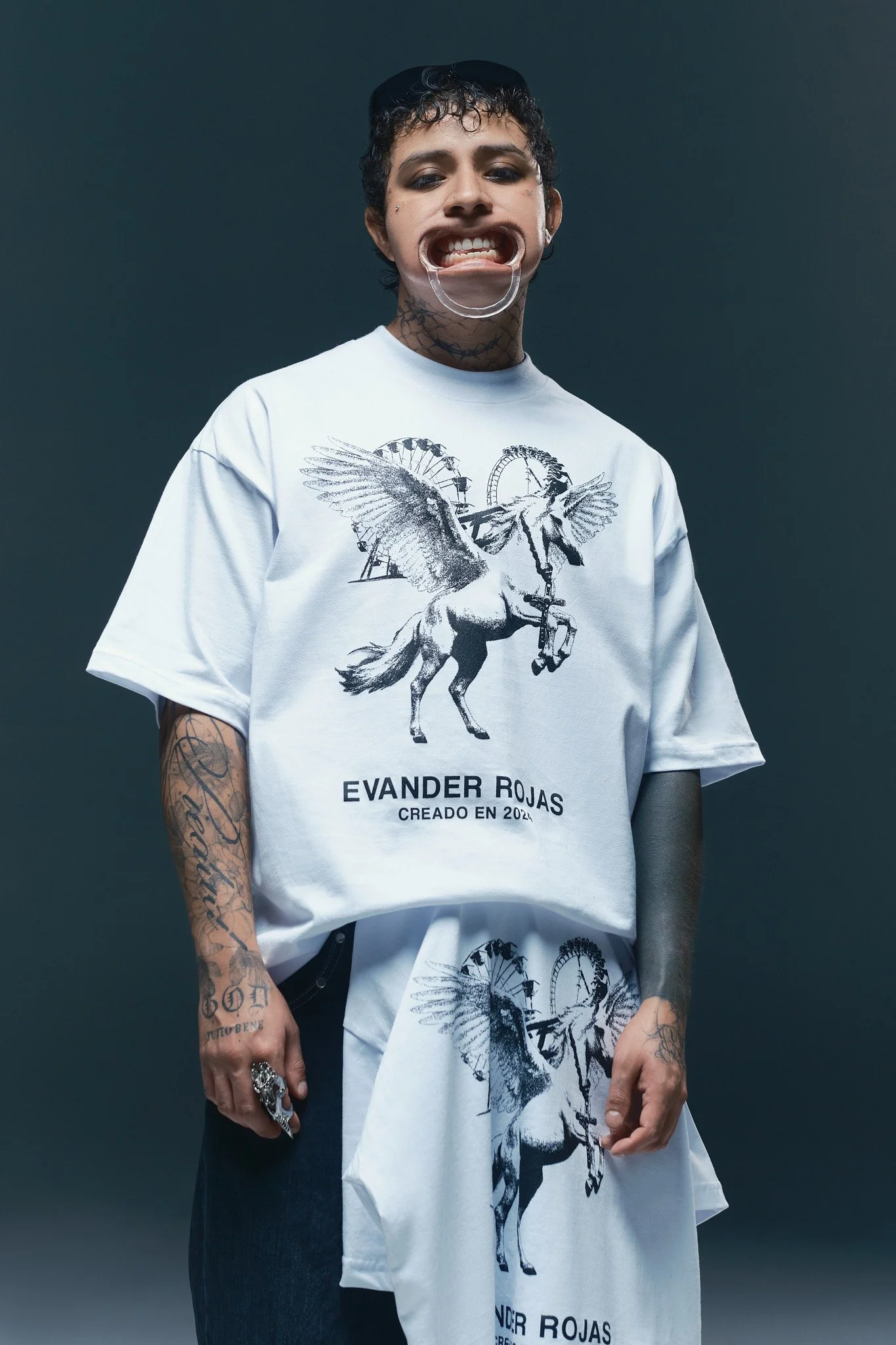 Joven con tatuajes en los brazos y cuello, viste una camiseta blanca con estampado de caballito con alas y un globo de feria, y una gorra negra, posando con expresión audaz sobre fondo gris oscuro.