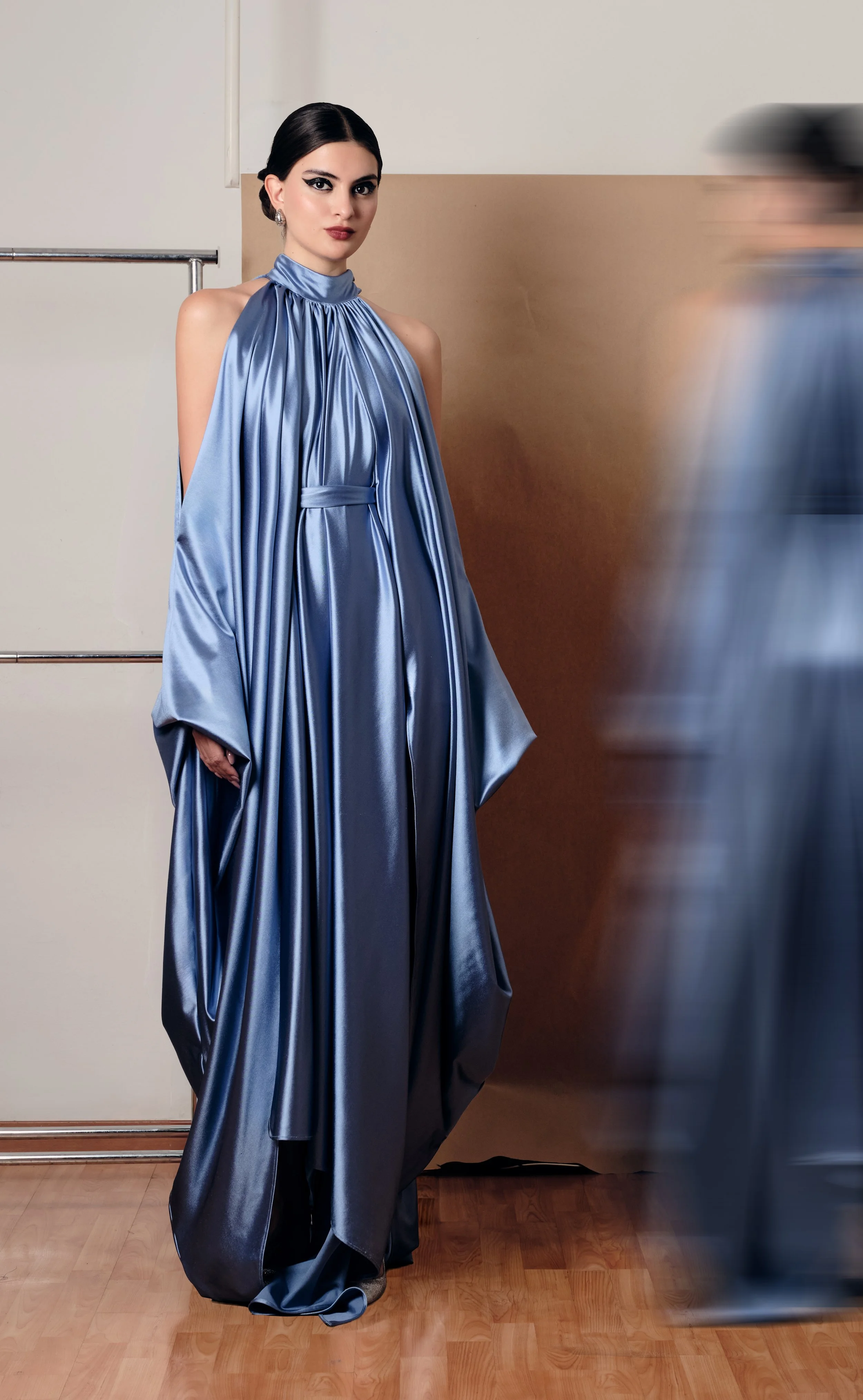 Mujer con vestido largo y satinado de color azul, con el cabello recogido, de pie en un interior, mientras otra persona pasa rápidamente a su lado.