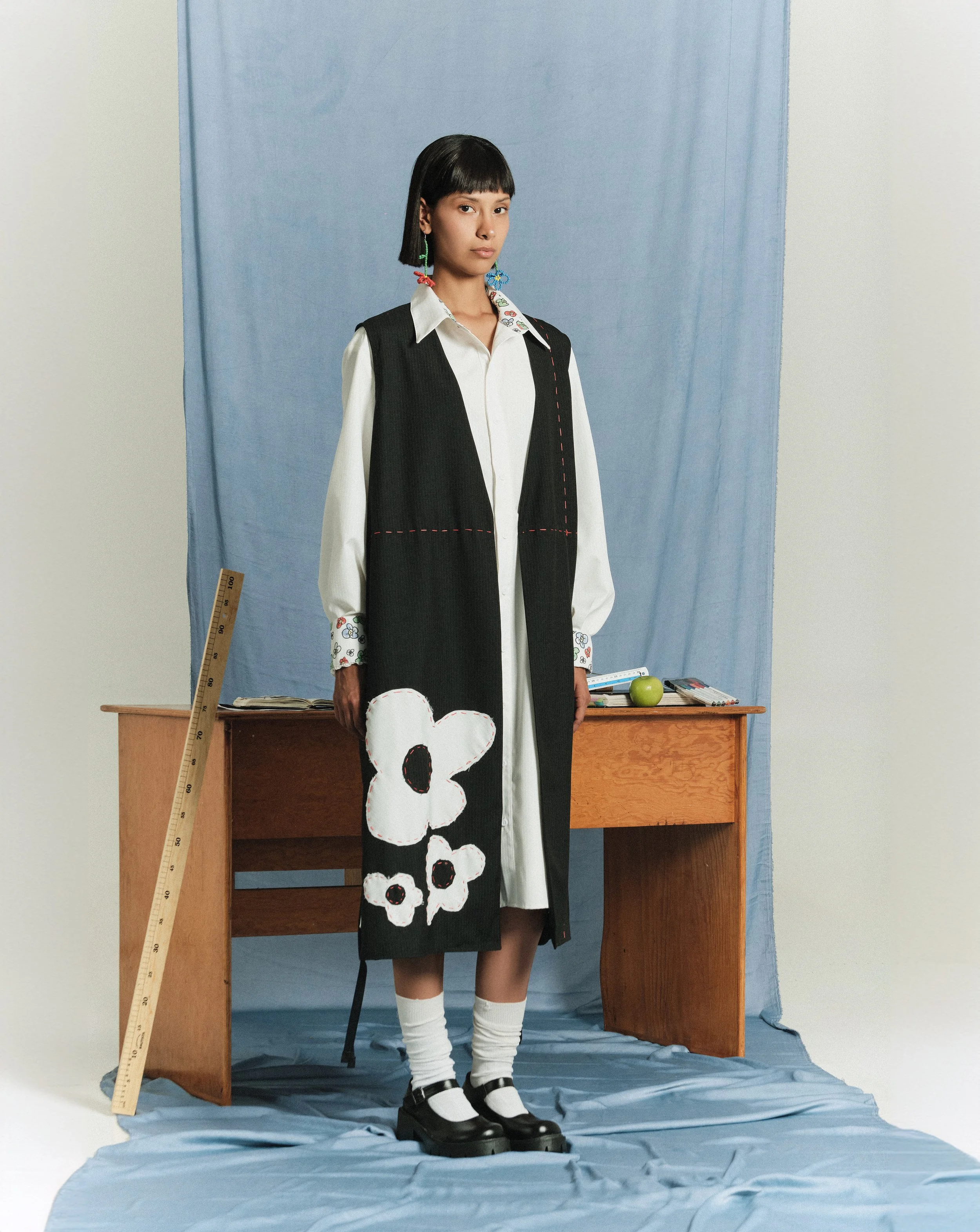 Mujer con vestido largo decorado con flor blanca en la pierna, camisa blanca, chaleco negro, zapatos negros y calcetas blancas, en un estudio con fondo azul y mesa de madera con objetos diversos.