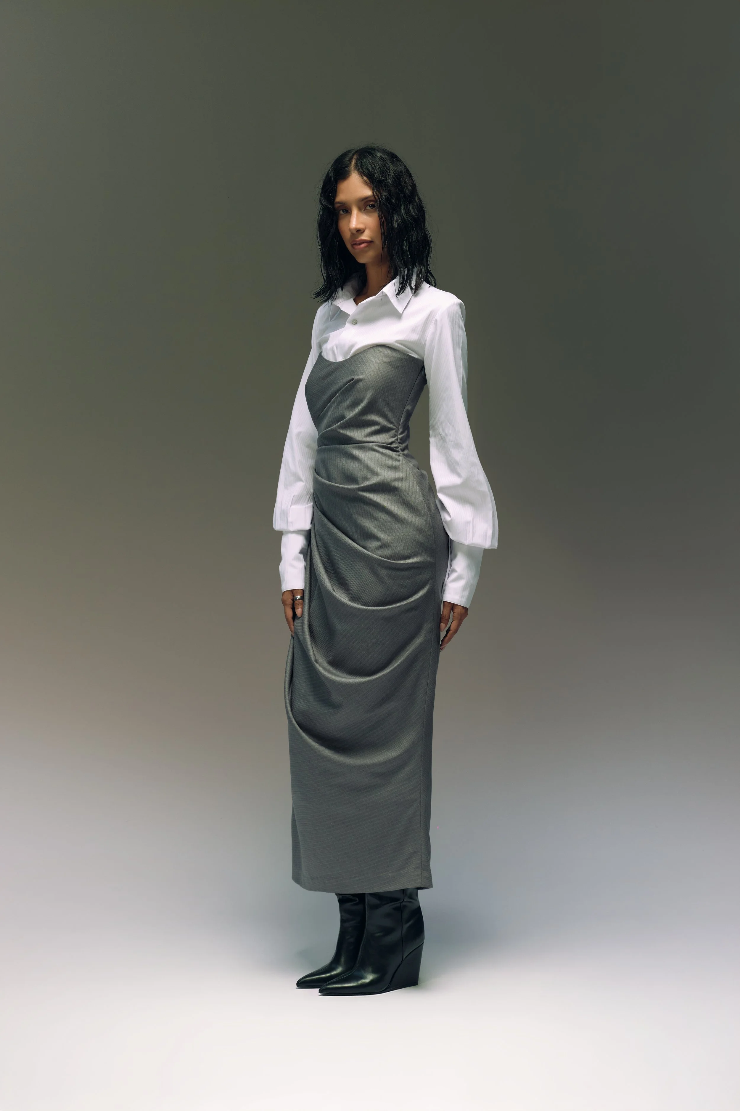 Mujer con vestido largo gris y blusa blanca con mangas largas, usa botas negras de tacón alto, de pie con fondo gris.