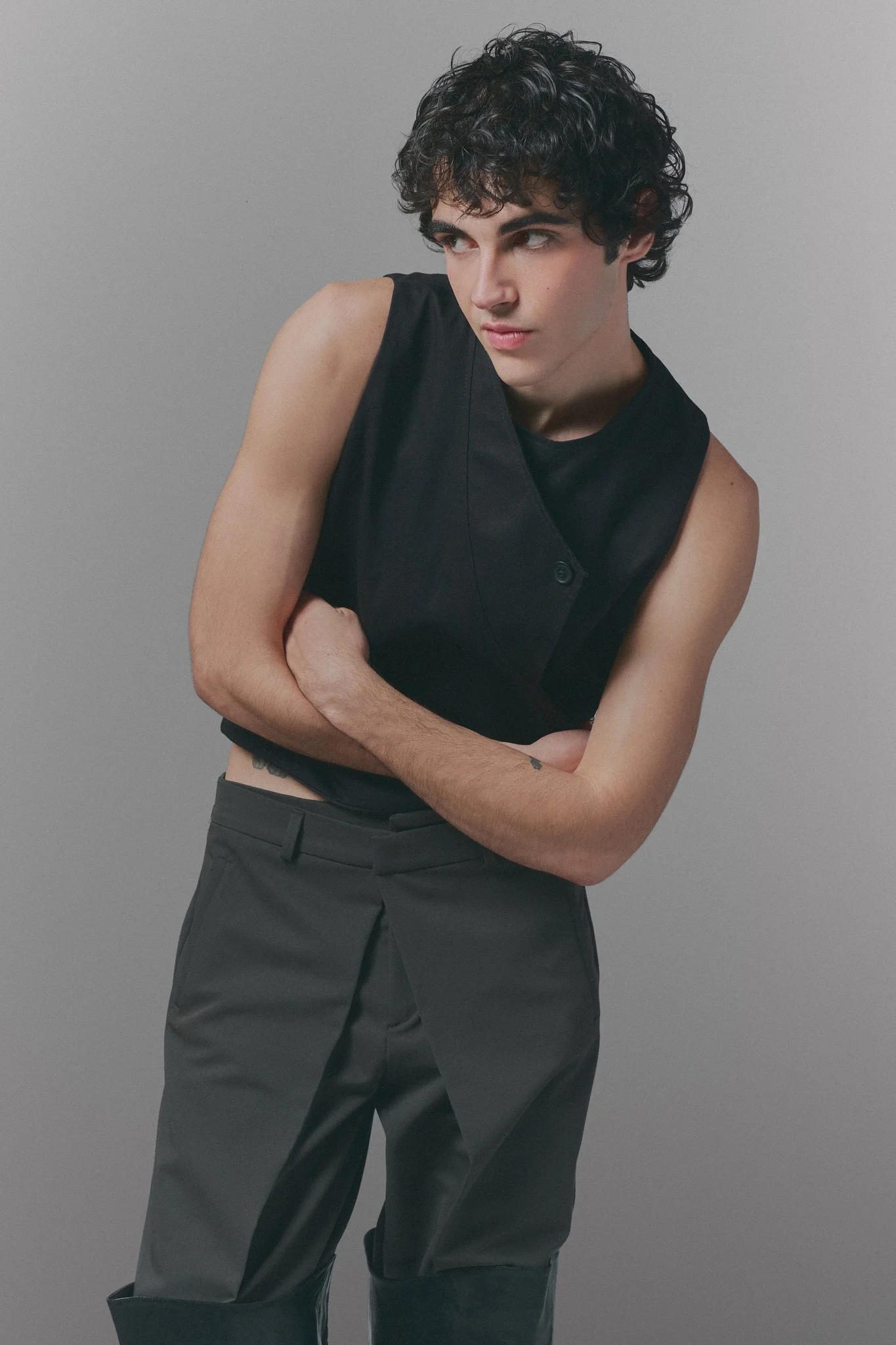 Joven de cabello rizado y oscuro, vestido con pantalones oscuros y sin camisa en un fondo gris, mirando hacia la izquierda con expresión pensativa.