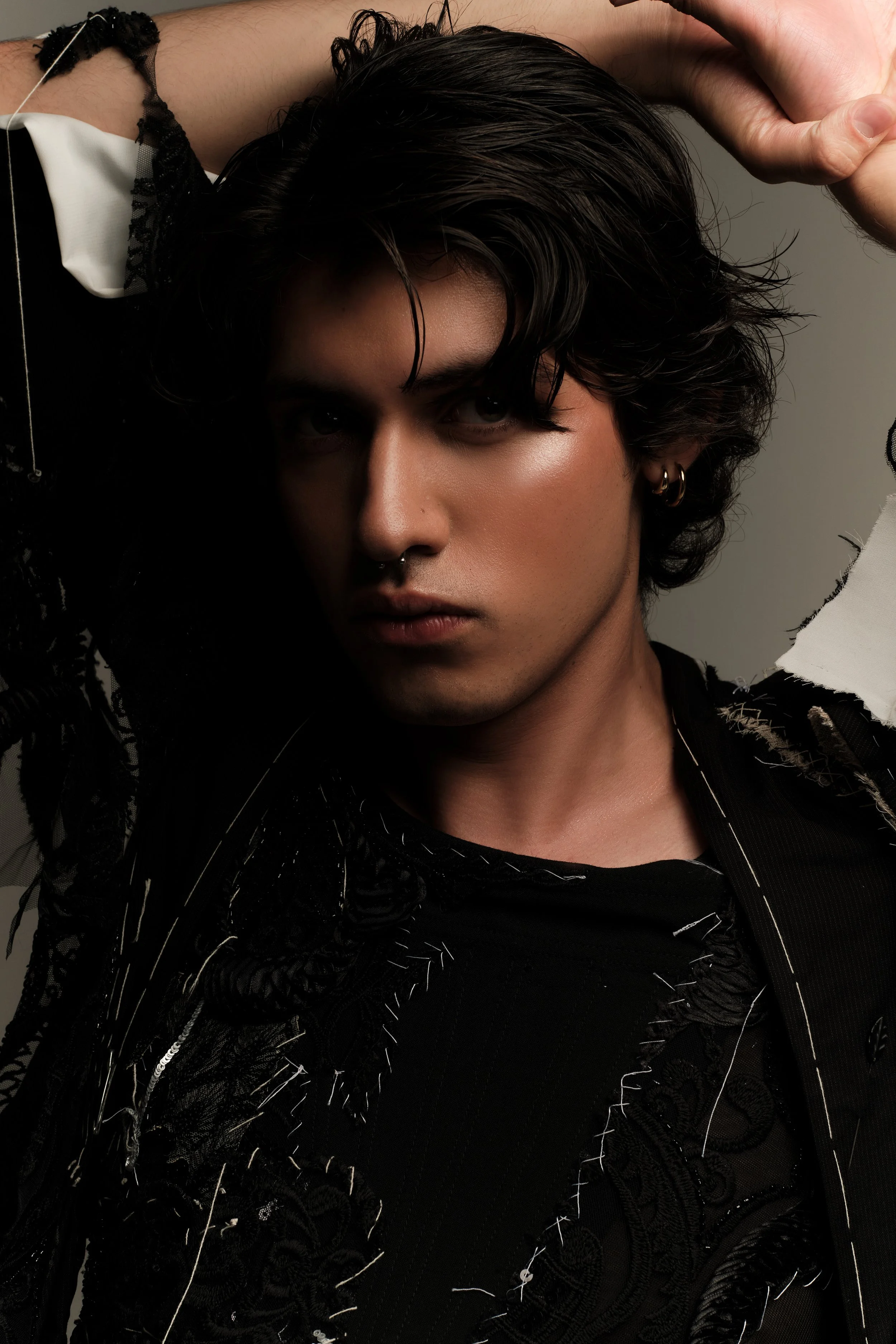 Retrato de un joven con cabello oscuro y largo, usando prendas negras y con accesorios en las orejas, en un fondo gris oscuro.