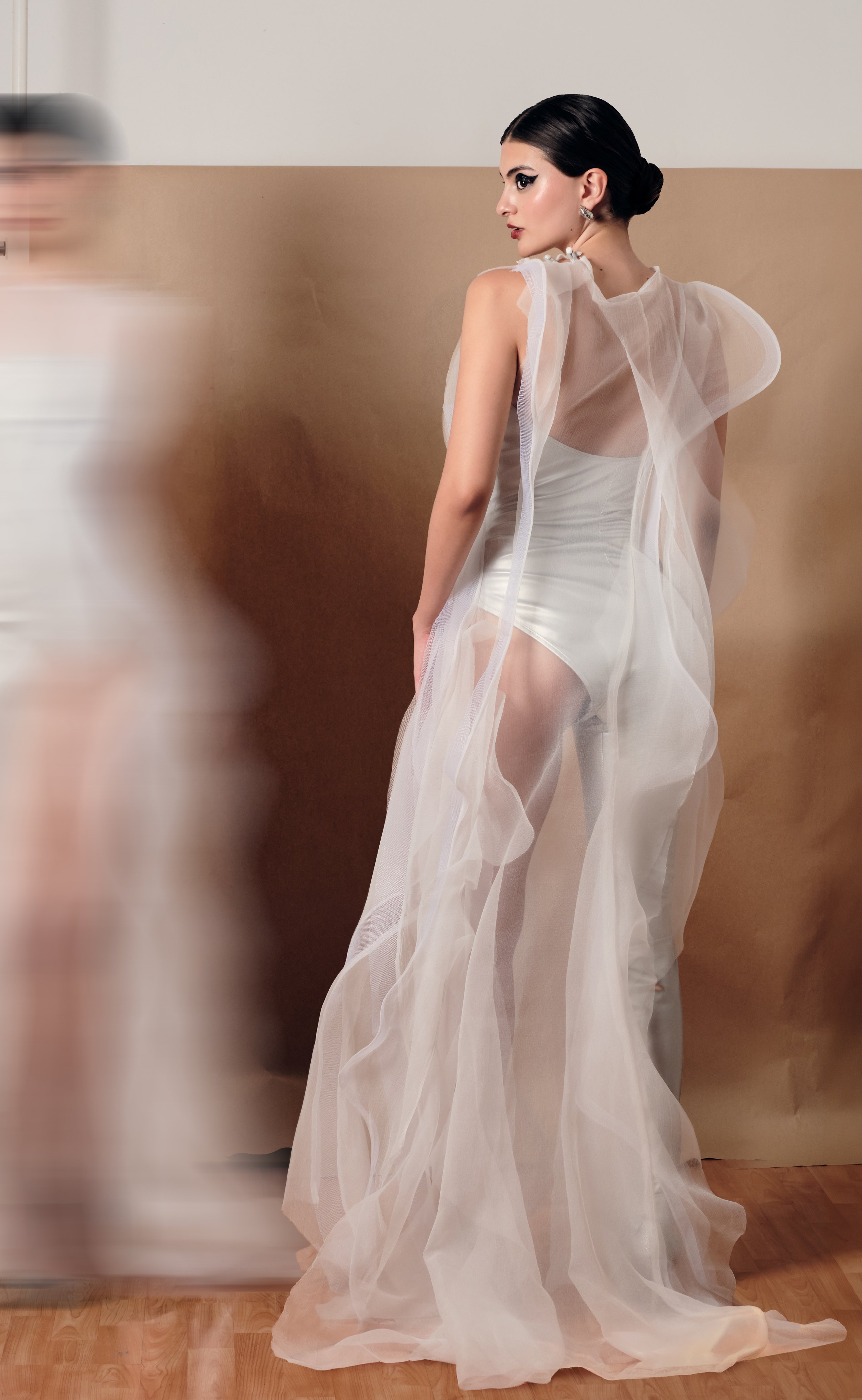 Mujer con peinado elegante, vestido blanco con tela transparente y forma vaporosa, de perfil, en un entorno interior.