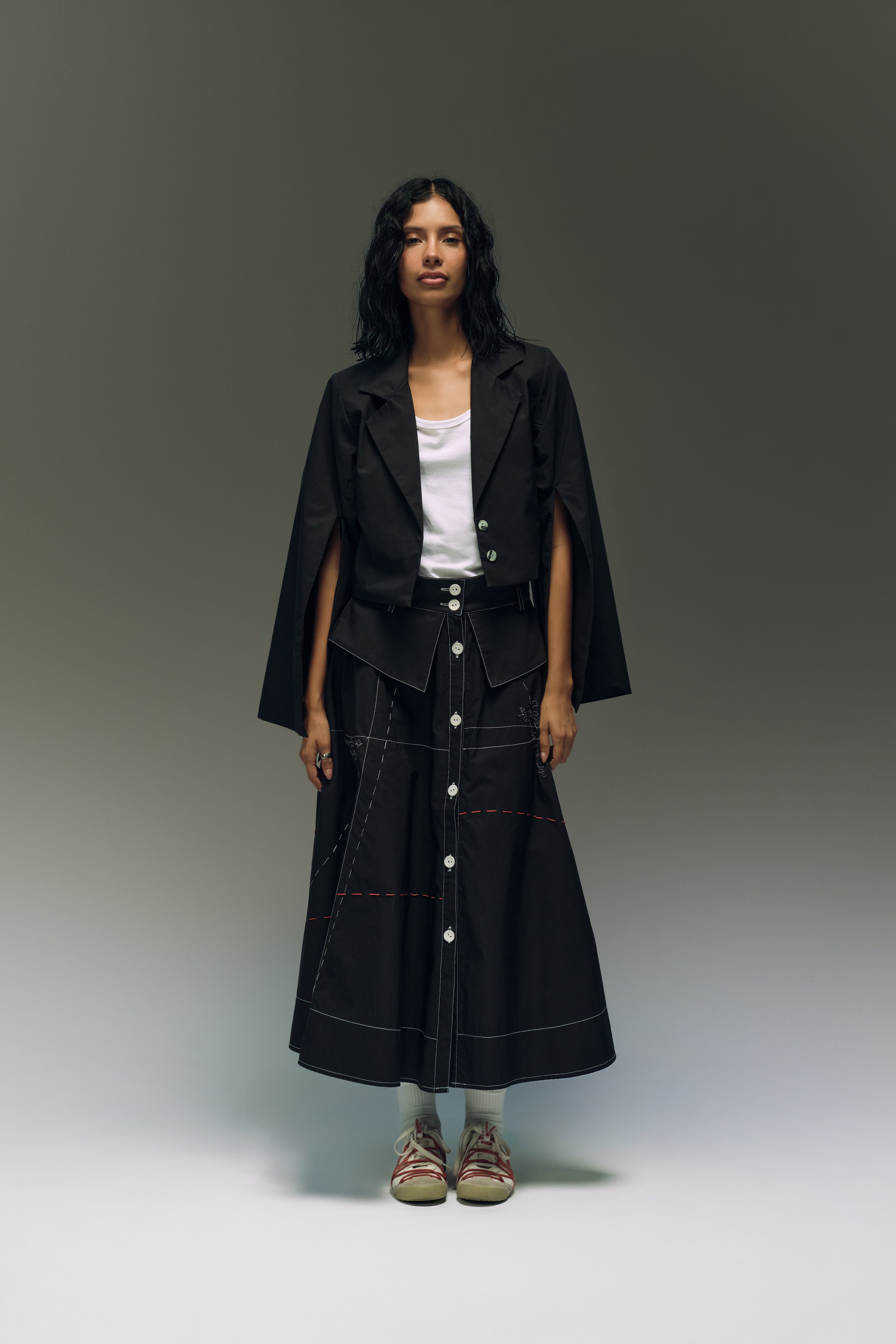 Una mujer con cabello rizado negro, ropa moderna y fondo gris.