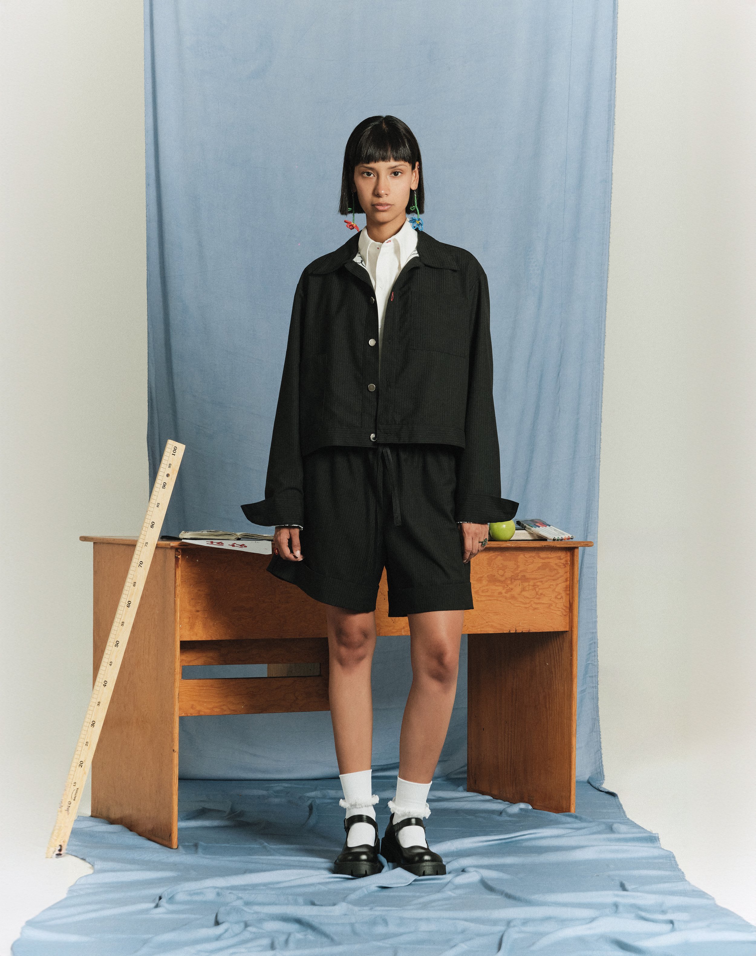 Una joven viste ropa oscura y calcetas blancas con zapatos negros, se encuentra de pie frente a un escritorio de madera con objetos en la superficie, detrás de ella hay una cortina azul y un fondo blanco, en un entorno de estudio o retrato.