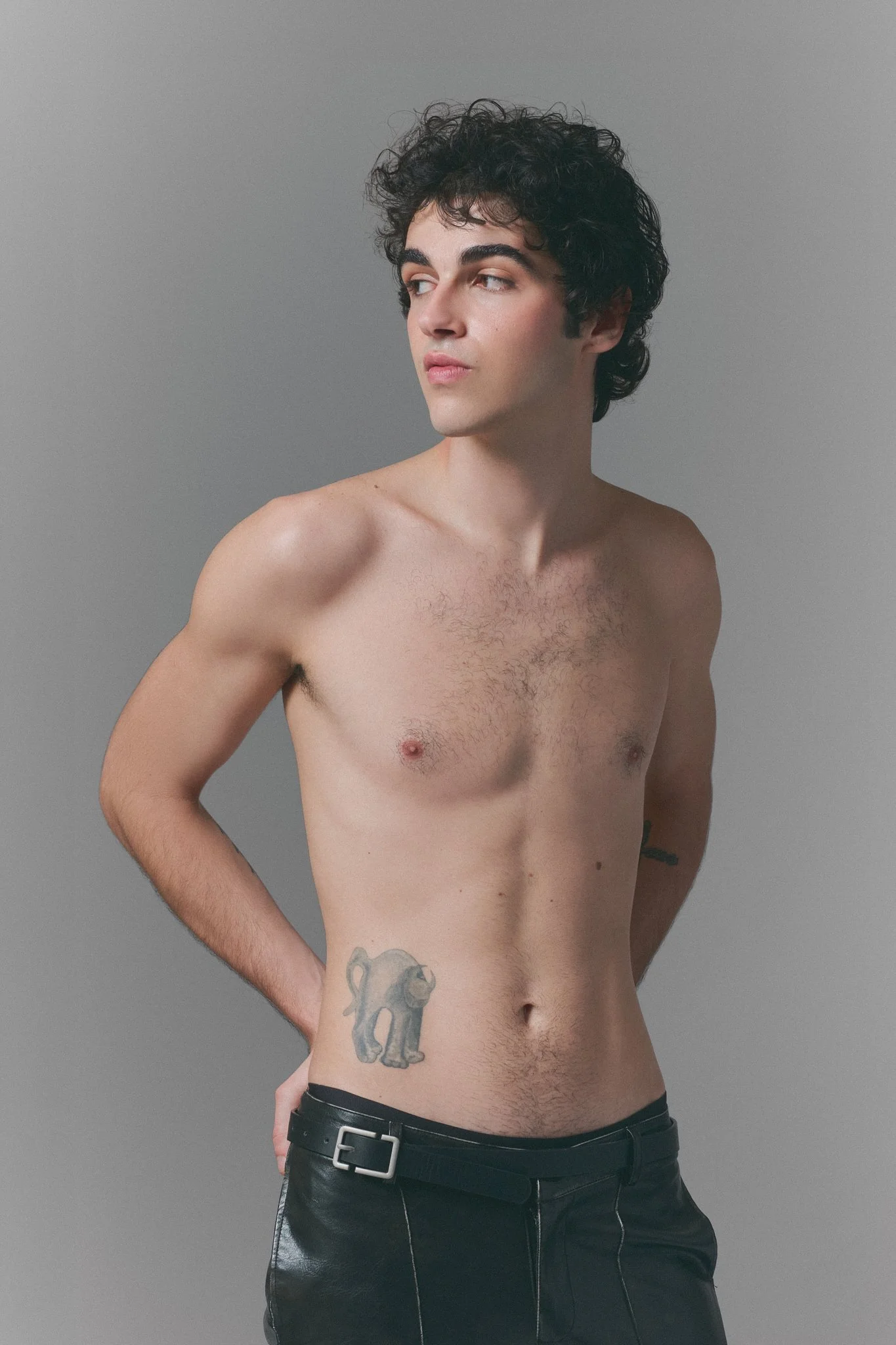 Joven desnudo de género masculino con tatuaje de elefante en el lado izquierdo del abdomen, de cabello rizado y oscuro, con pantalones negros, posando de perfil contra fondo gris.