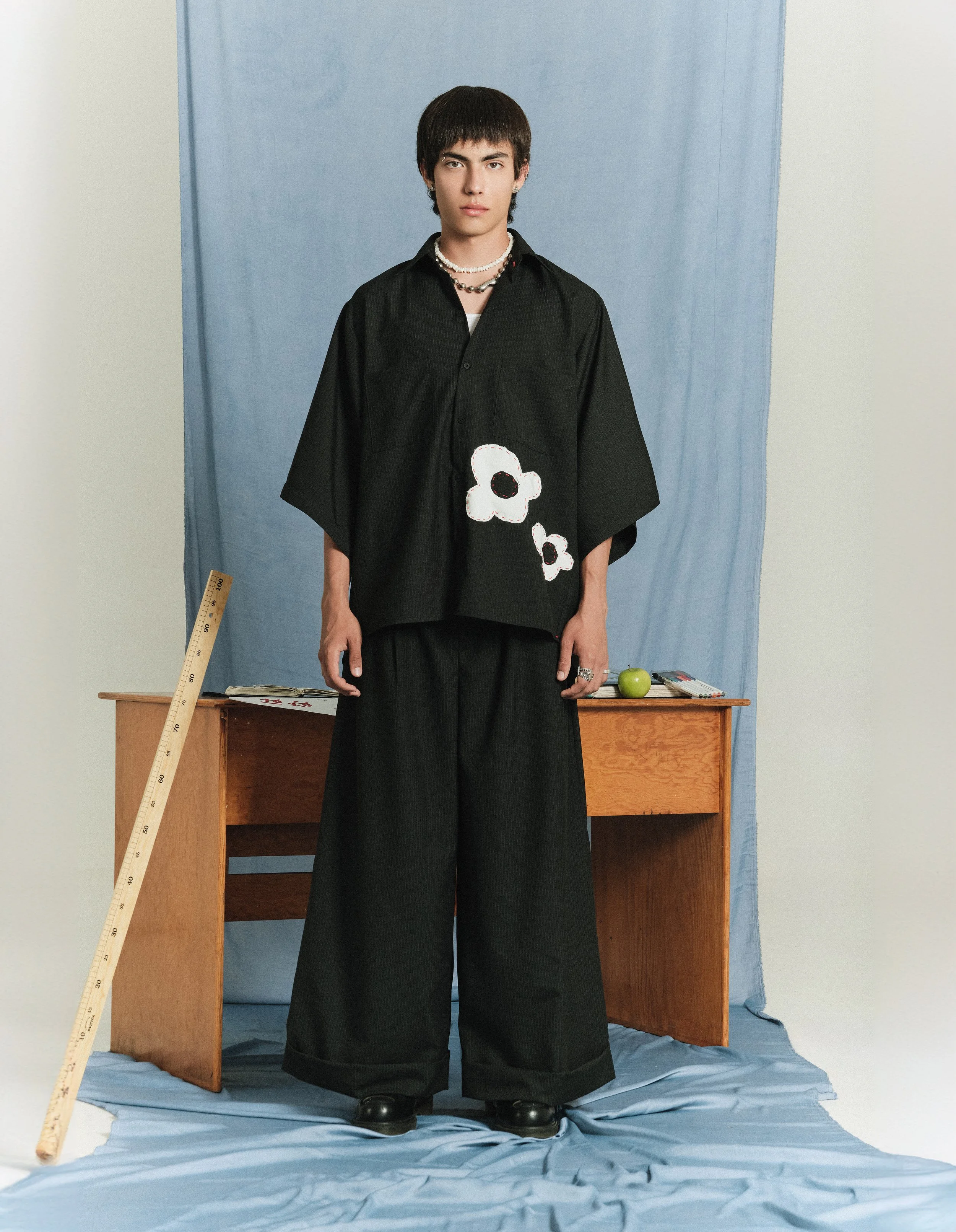 Joven con ropa negra con detalles de dibujos de flores, de pie frente a fondo azul y un escritorio de madera con una regla, manzana verde y libros.