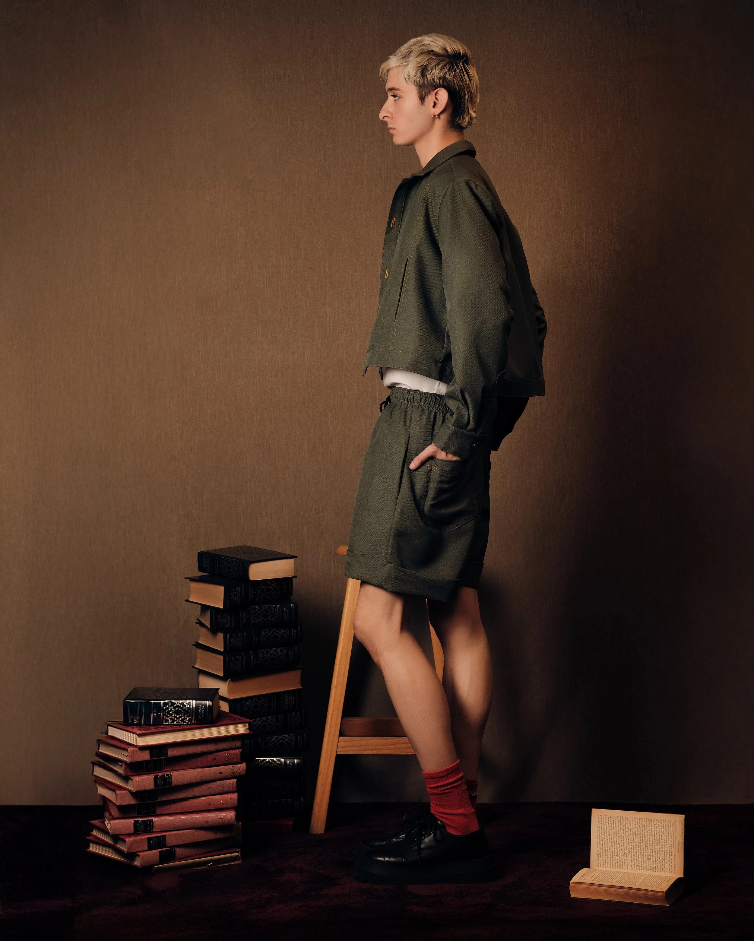 Joven con cabello rubio, con pantalones cortos y chaqueta, de perfil, rodeado de pilas de libros, uno abierto en el suelo, en un fondo liso y oscuro.