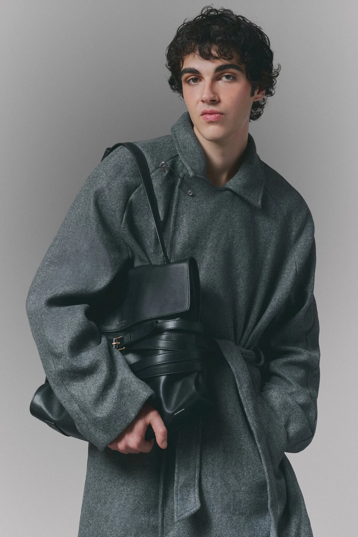Joven con cabello rizado y oscuro, vestido con abrigo gris y cinturón negro, cargando una bolsa negra, posando de perfil con fondo gris.