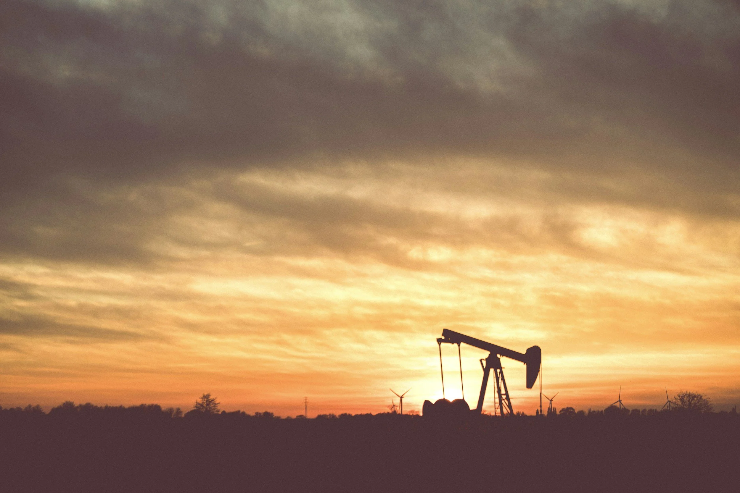 Silhouetted oil pump jack against a sunset sky with wind turbines in the background.