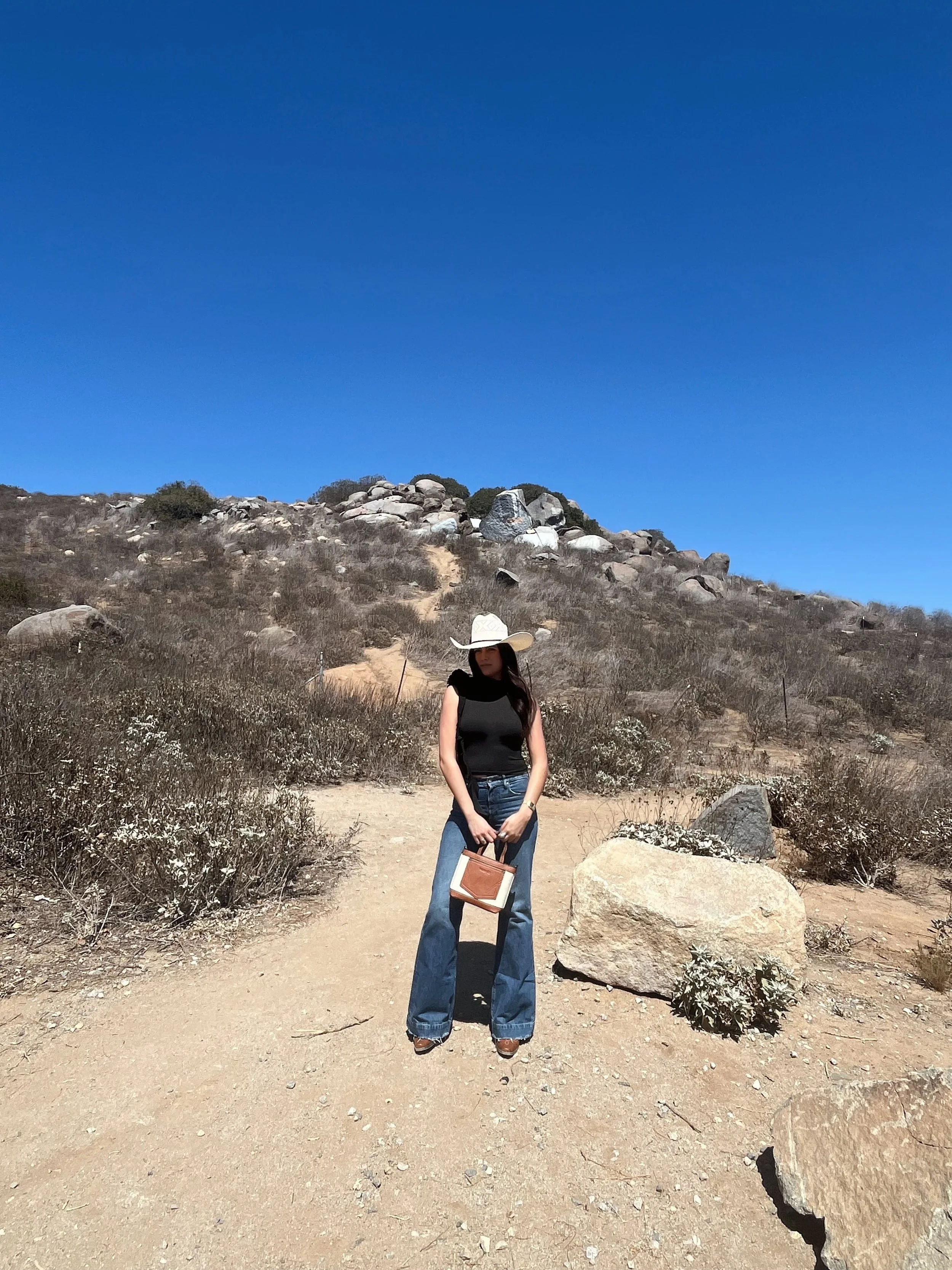 Woman standing on dirt trail in a desert landscape, wearing a wide-brimmed hat, black sleeveless top, blue jeans, holding a small handbag, with rocky hill and clear blue sky in background.