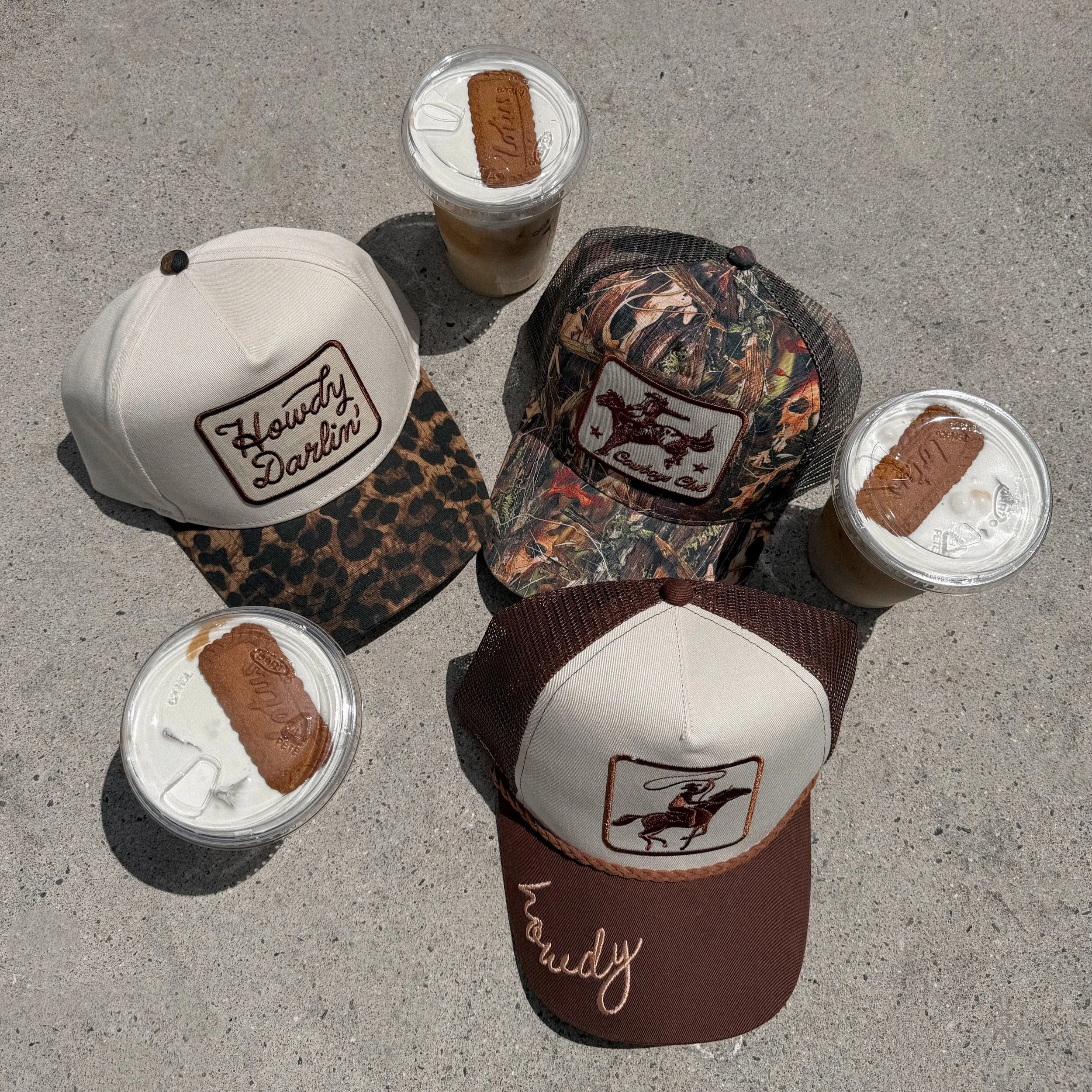 Three baseball caps and three iced coffee drinks on a concrete surface. The caps have various designs, including leopard print, floral camouflage, and a horse logo. The iced coffee drinks have a cookie on top of the whipped cream.
