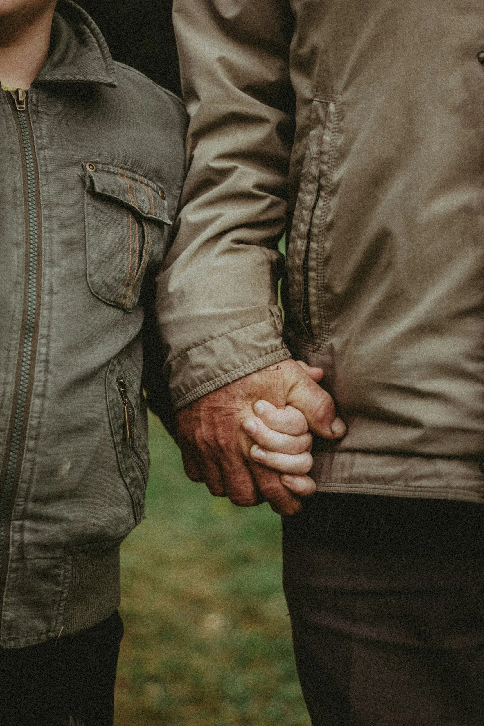 A close-up of two people holding hands, one appears to be older with visibly aged skin, and the other is younger, both dressed in casual jackets.