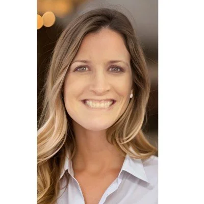 A smiling woman with long wavy hair wearing a white collared shirt, with blurred background lighting.
