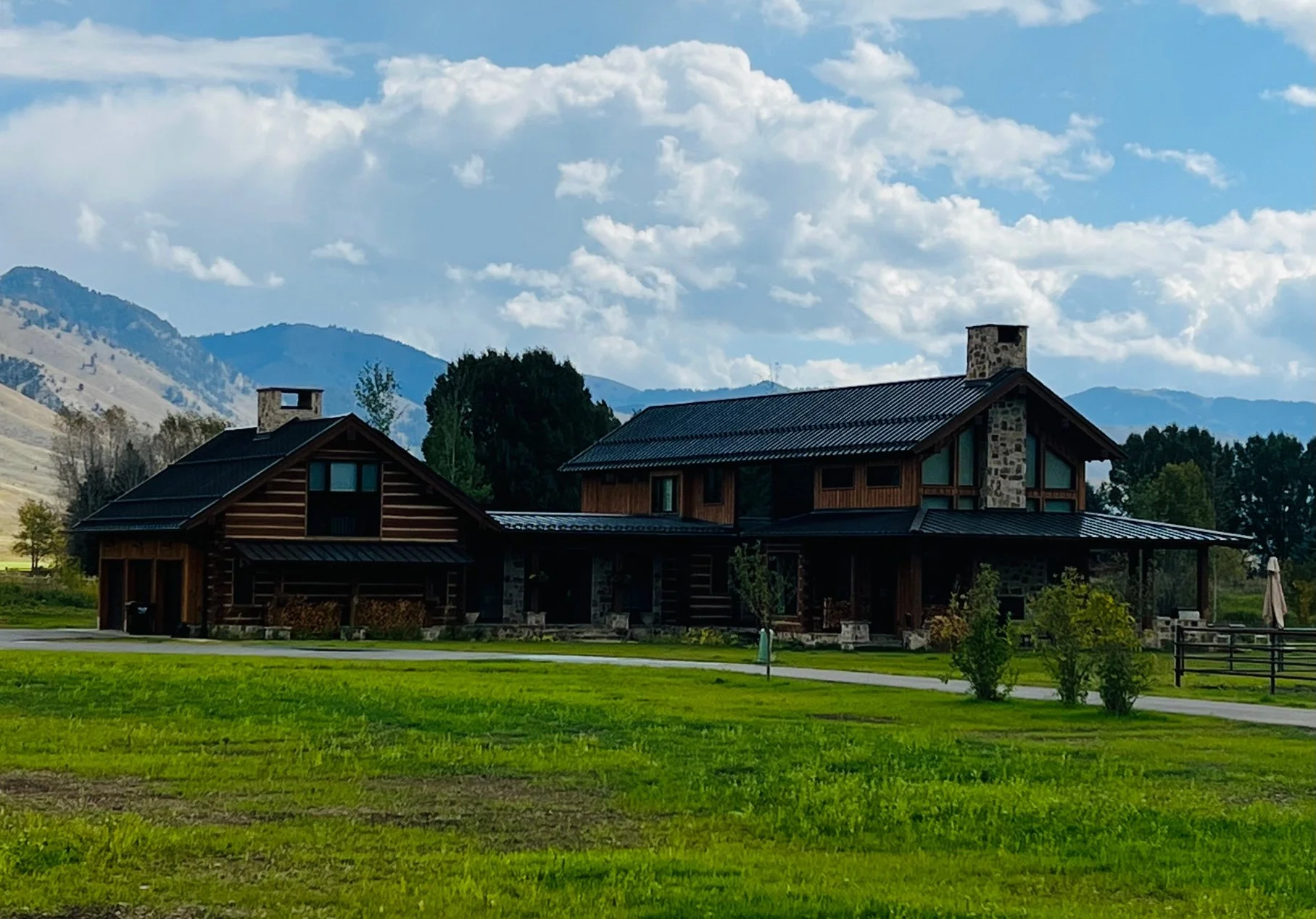 A large house with a dark metal roof and a stone chimney, situated in a grassy area with small trees, with mountains and a partly cloudy sky in the background.
