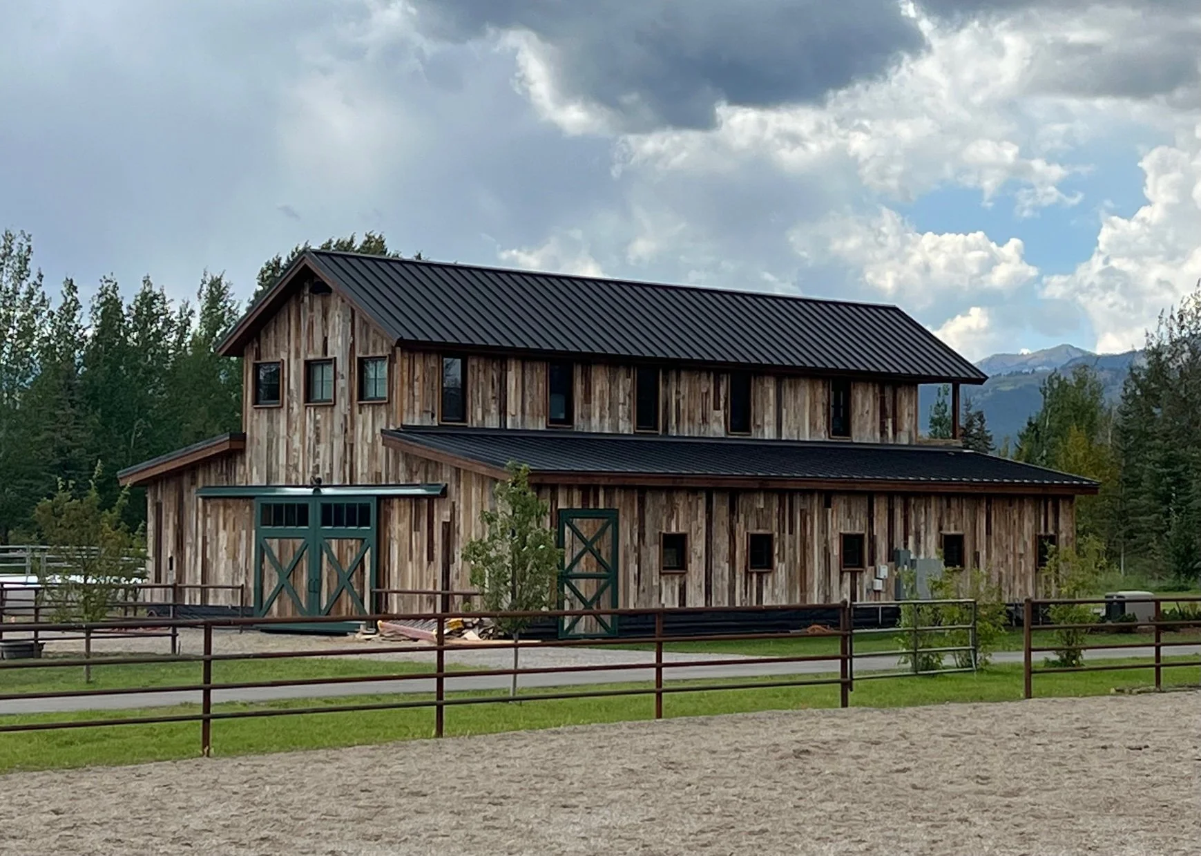 A rustic barn with wooden siding and a black metal roof, set against a backdrop of trees and mountains under a cloudy sky.