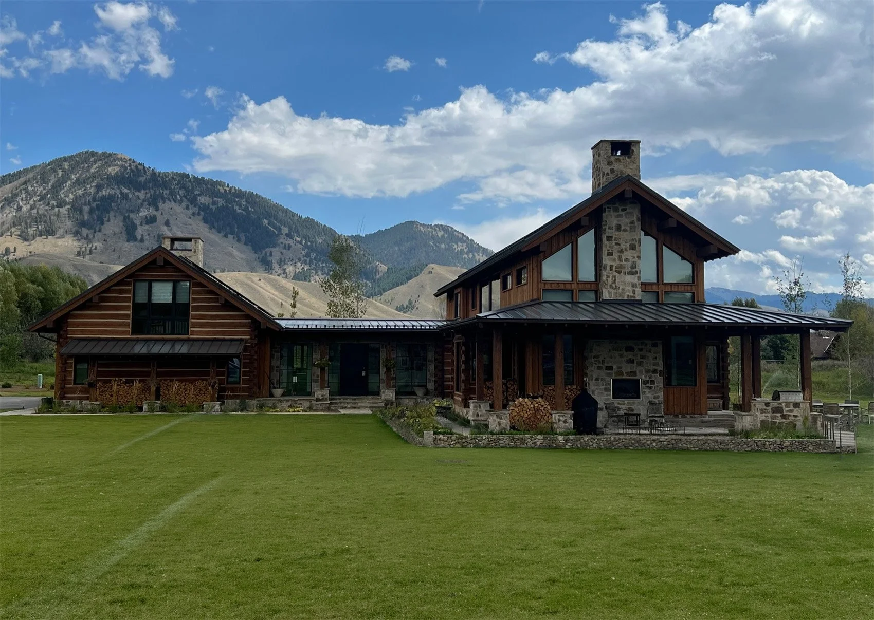 A large house made of wood and stone sits in a green field with mountains in the background. The house has multiple levels, large windows, and a metal roof, with stacks of firewood visible outside.