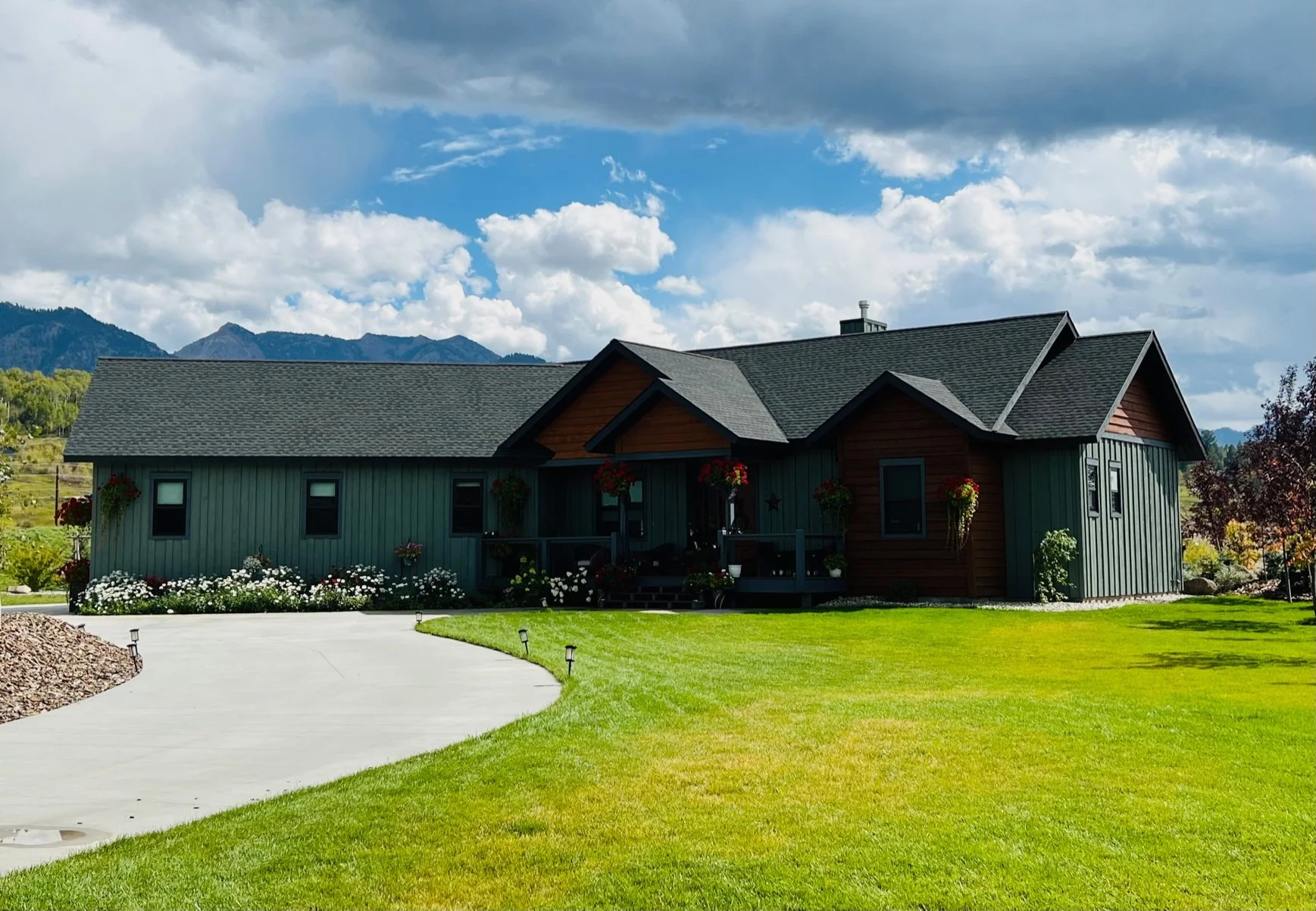 A house with green siding, brown accents, and a gray roof, situated in a lush green yard with a curved concrete driveway leading to it. The house features hanging flower baskets, front porch, and is set against mountains and a partly cloudy sky.