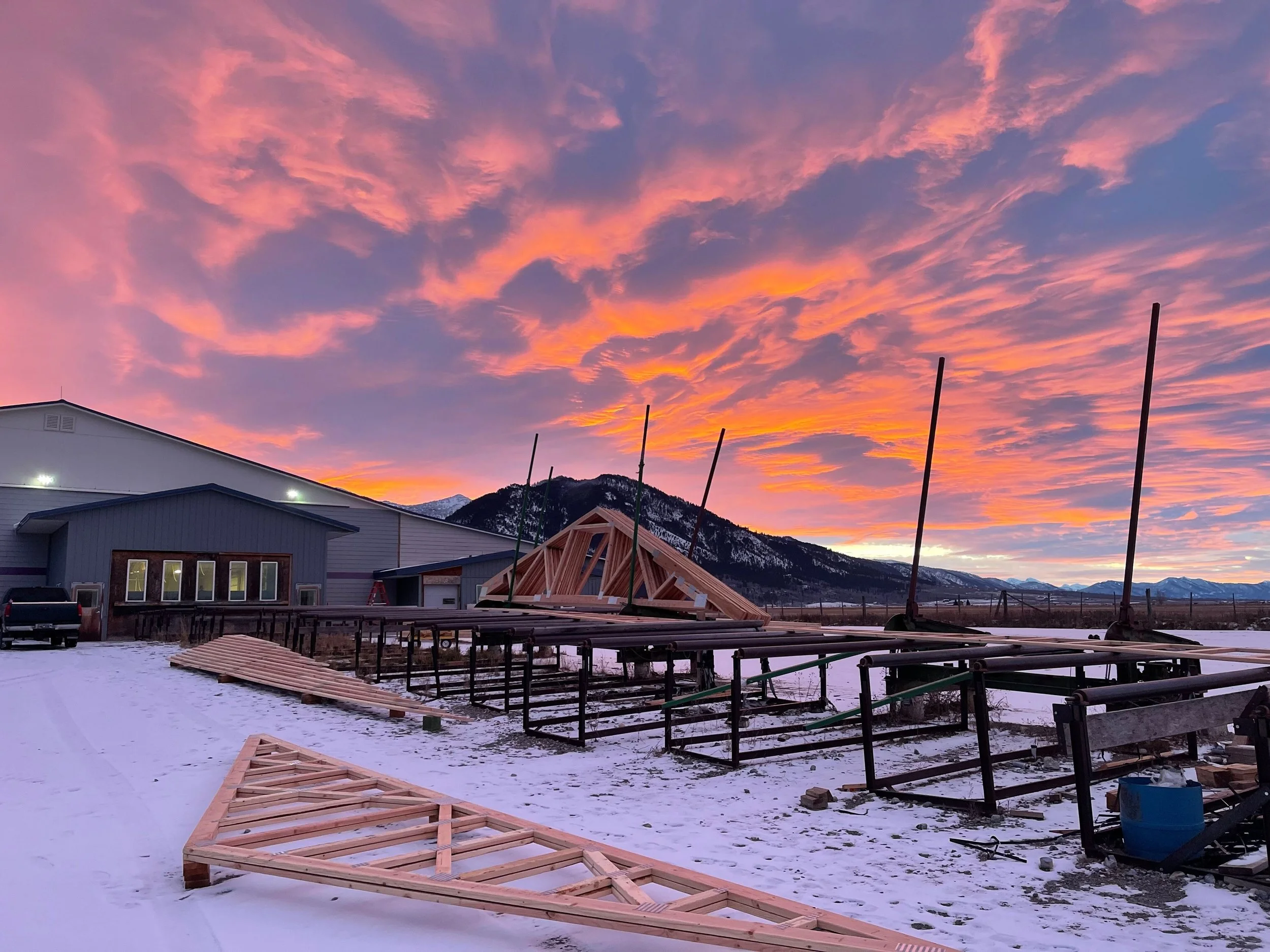 Construction site with wooden framing structures in snowy landscape during sunset, mountains in the background.