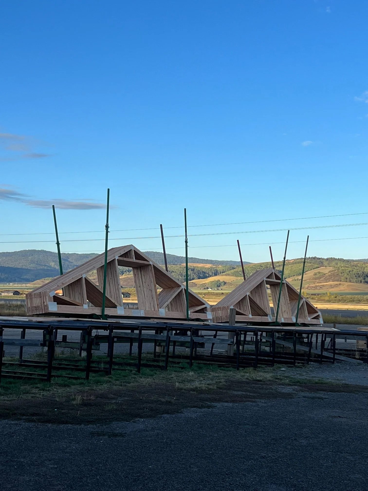 Wooden framed structures with construction poles set up around them, mountains and a bright blue sky in the background.