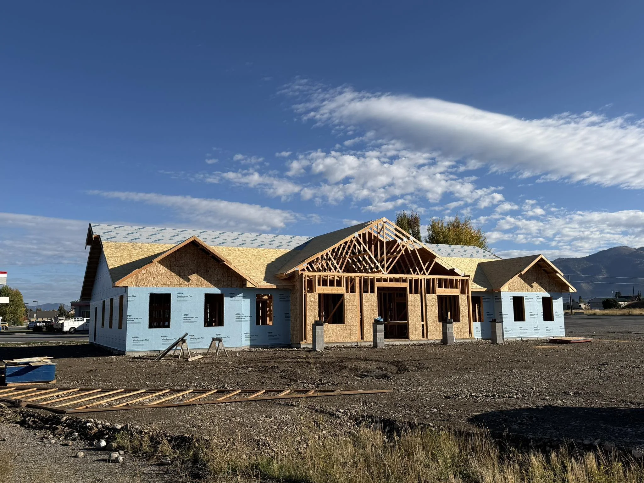 Under construction house with wooden framing and partly installed roof, set against a bright sunny sky with some clouds.