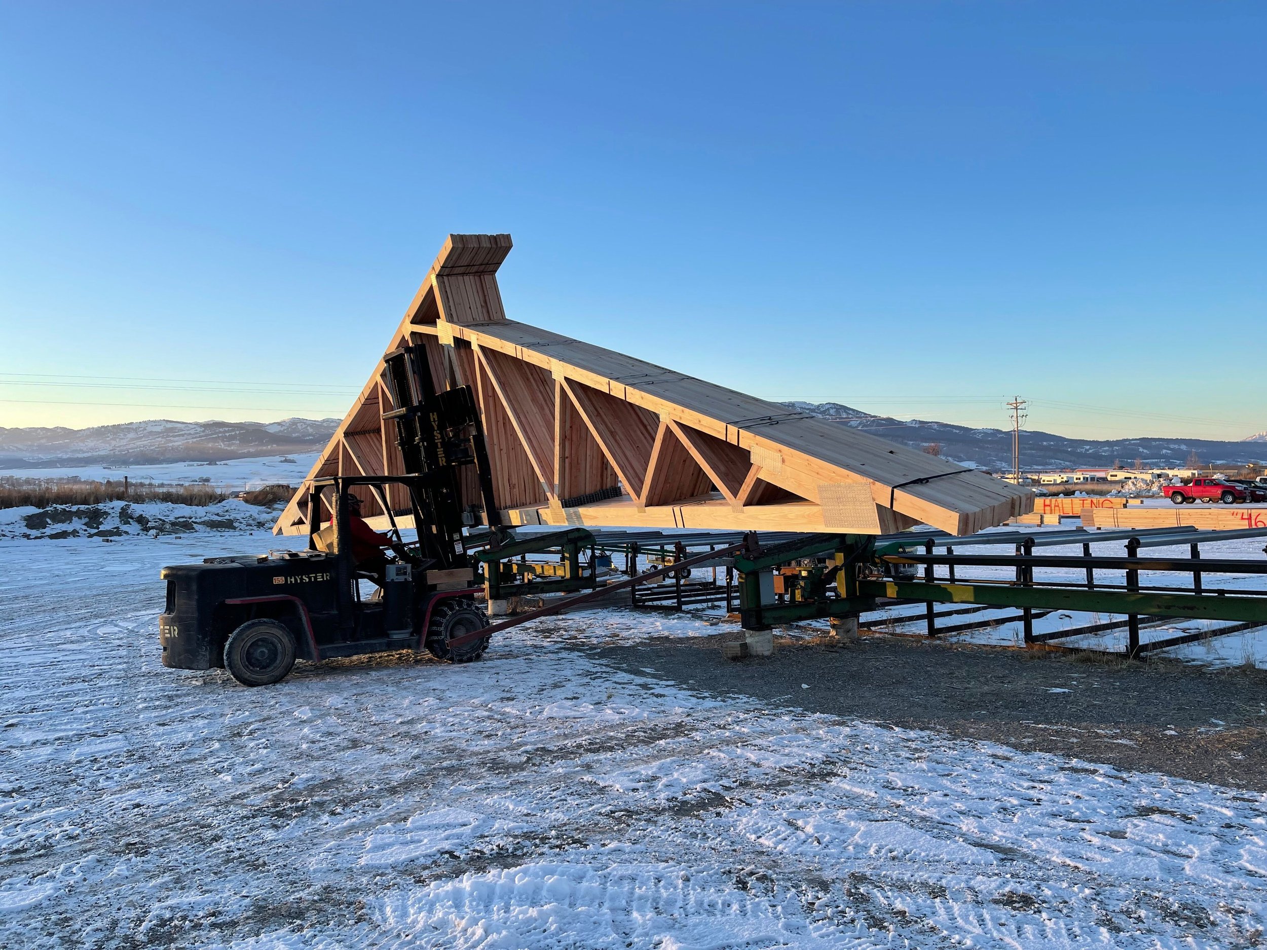 A large wooden structure under construction in a snowy outdoor area, with a forklift nearby and mountains in the background.