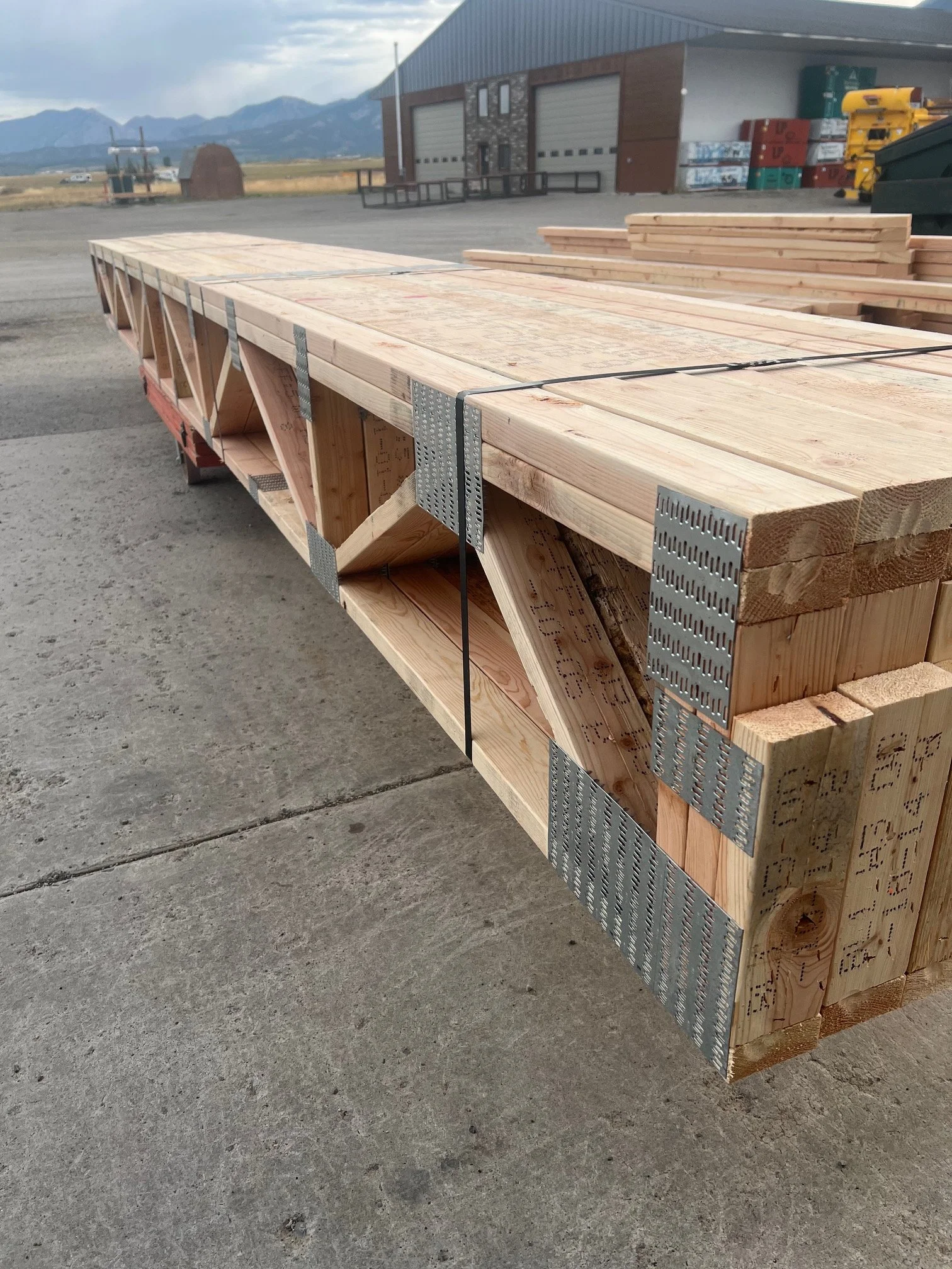 Stack of wooden planks and lumber on a flatbed trailer outside a warehouse, with mountains visible in the background.