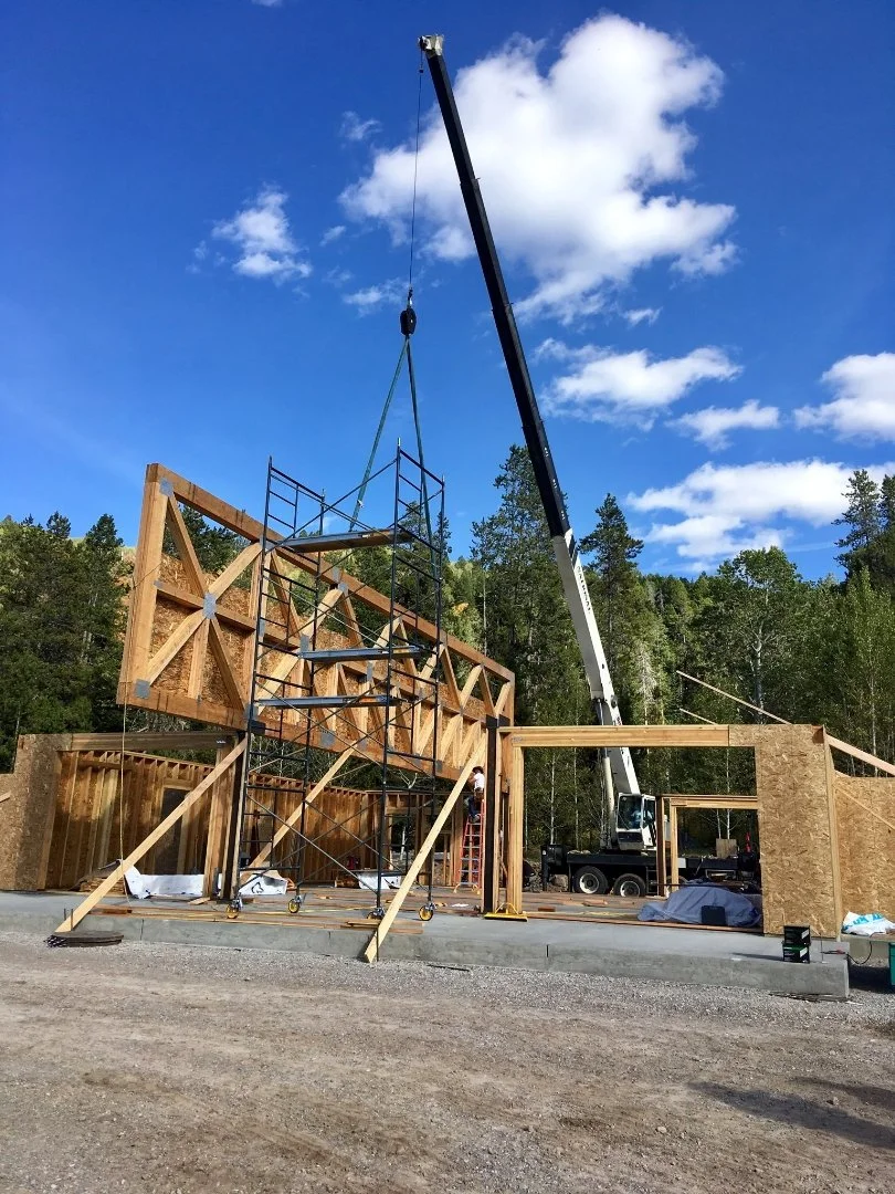 Construction site with a crane lifting wooden and metal framing for building walls, scaffolding set up, and a partly built structure against a backdrop of trees and a blue sky with clouds.