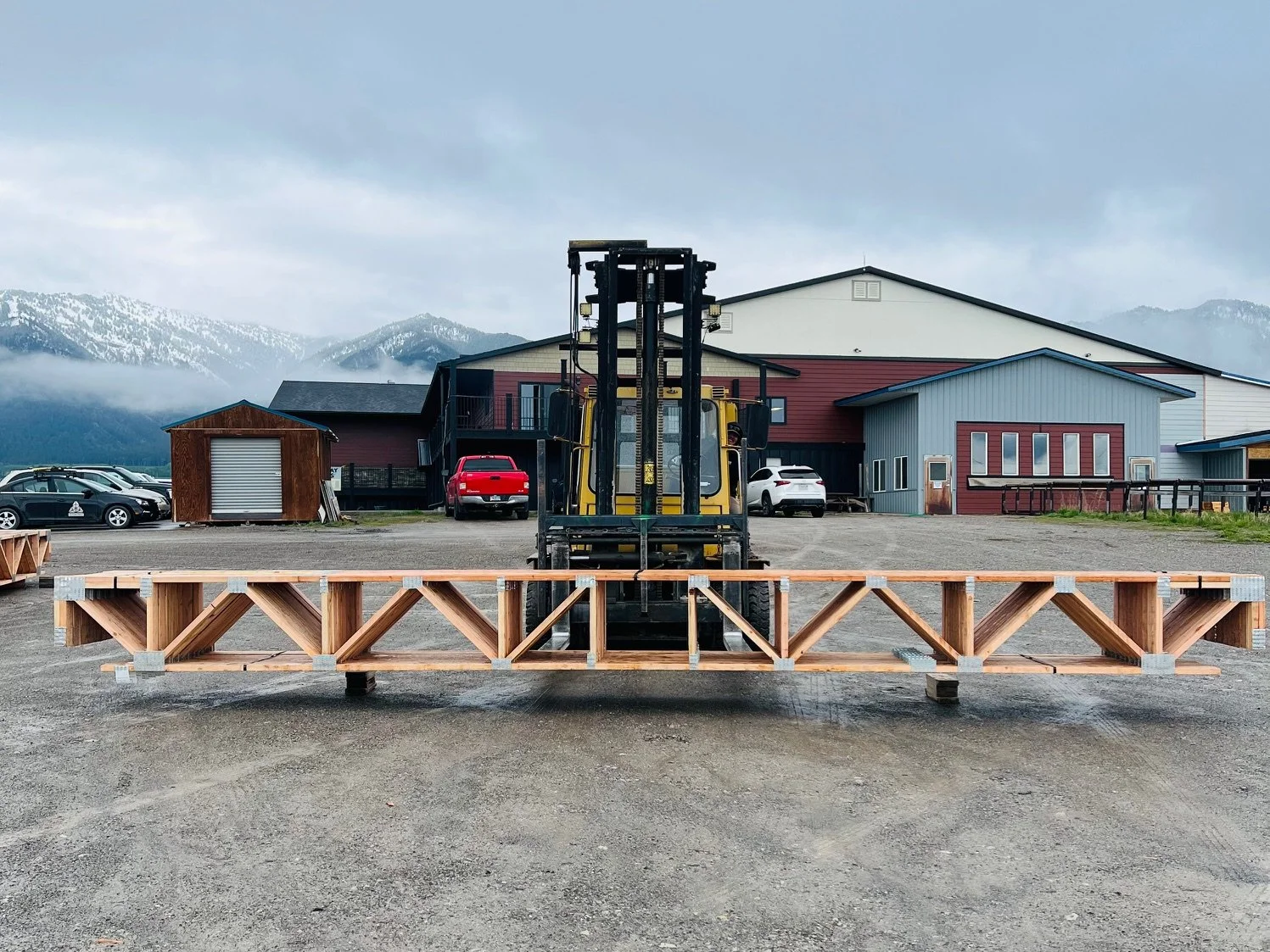 A forklift carrying a wooden structure parked in an open lot with multi-colored buildings and mountains in the background.