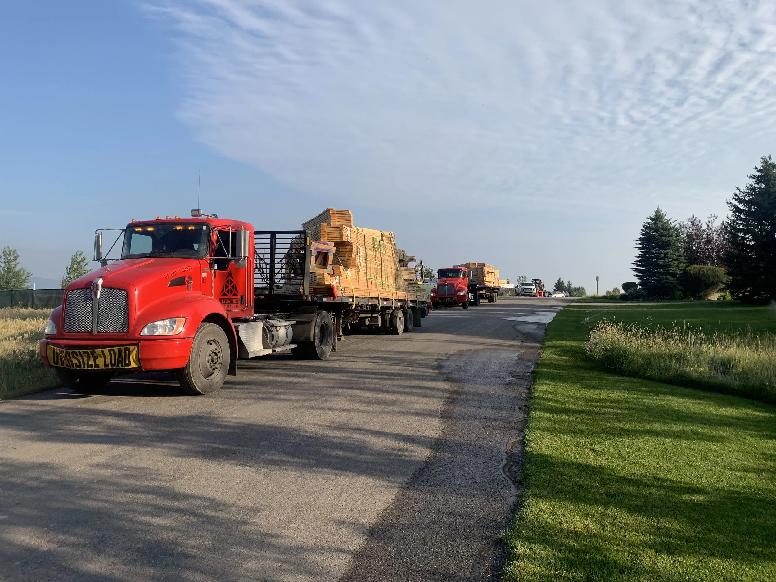 A red semi-truck carrying a load of wooden boards on a two-lane road, with a grassy area and trees on the side, under a partly cloudy sky.