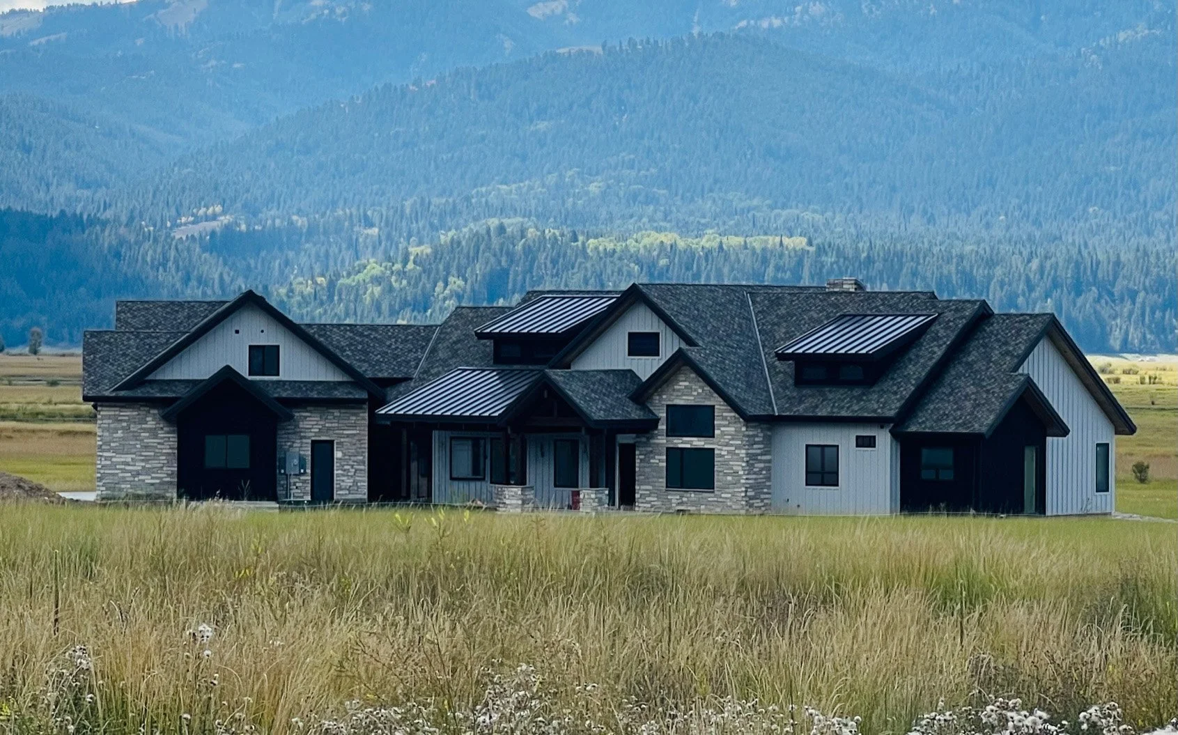 A modern house with gray siding and stone accents, situated in a grassy field with mountain in the background.