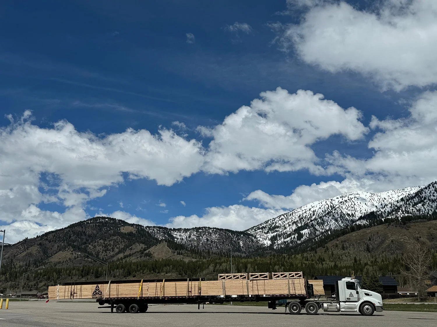 A flatbed truck with wooden panels driving along a road in front of a mountain range with snow-capped peaks and a partly cloudy sky.