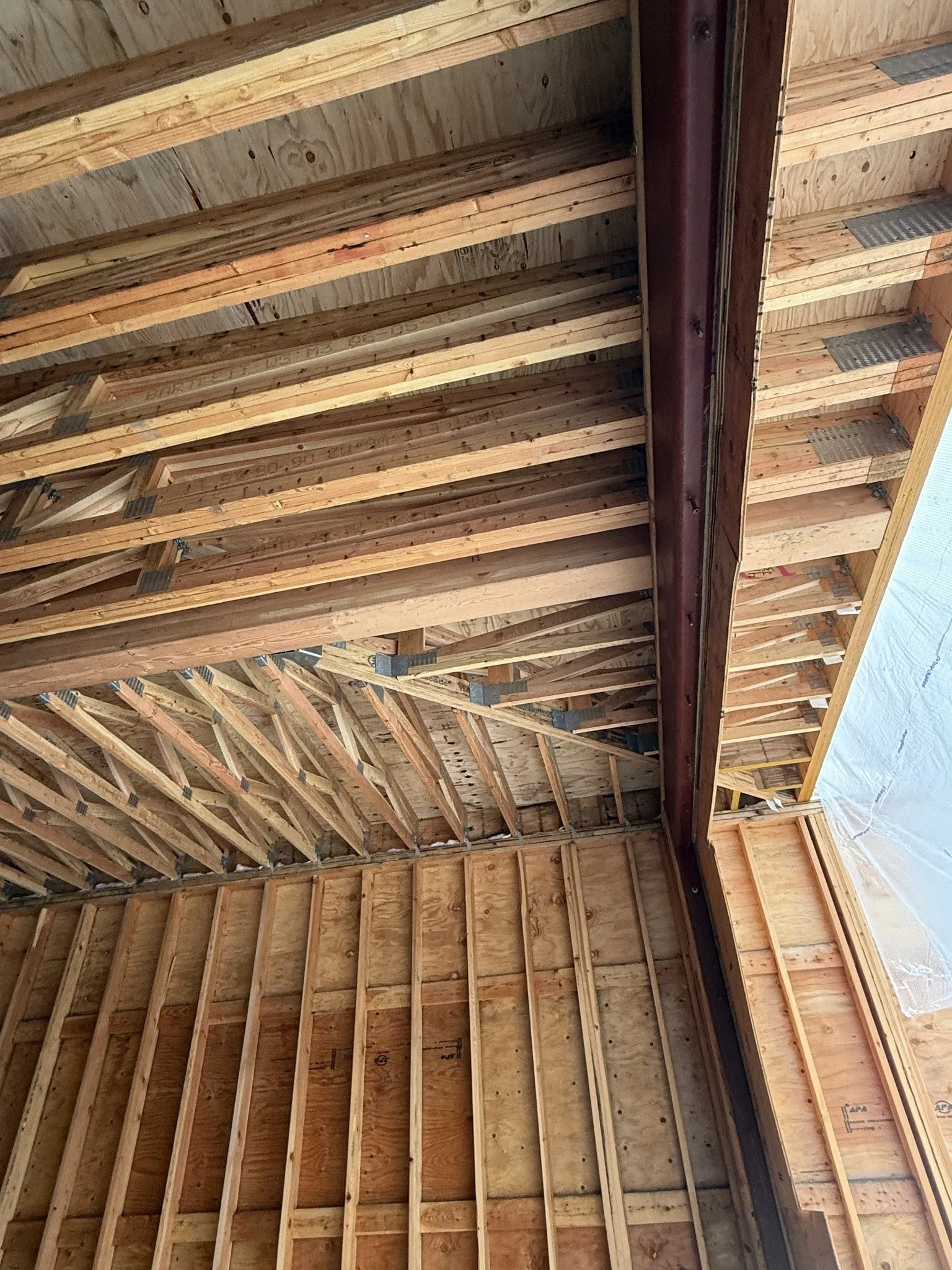 Interior view of a house under construction, showing wooden framing for the ceiling and walls, with some metal brackets and a metal pipe or beam.
