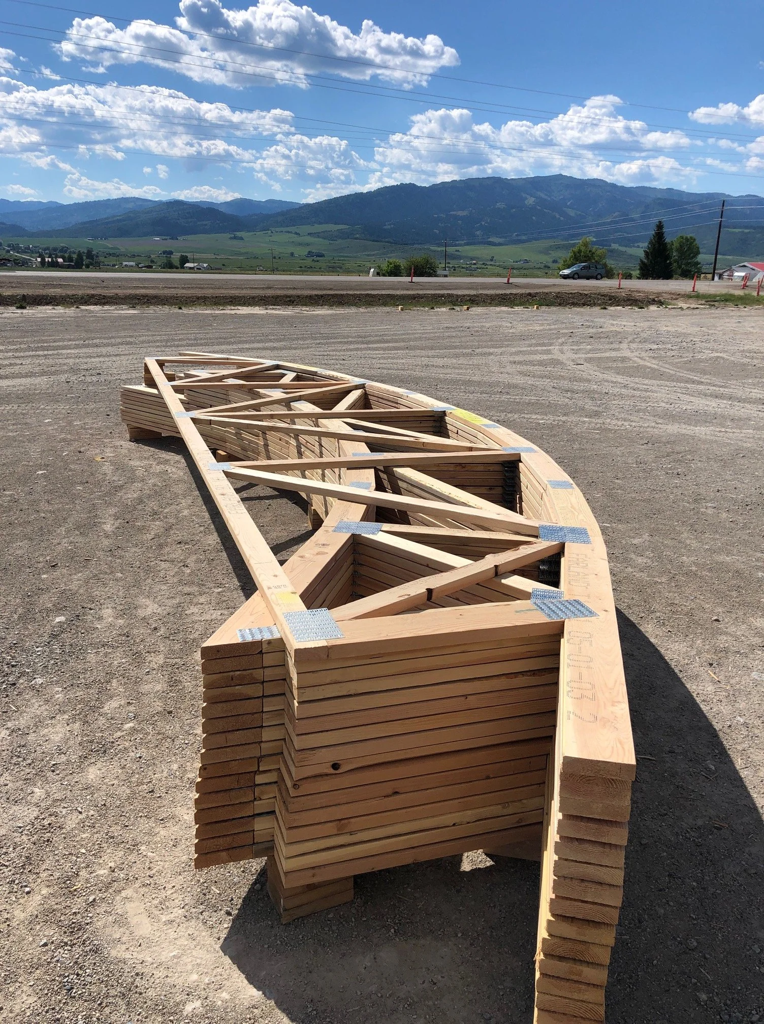 Stacked wooden boat frames on a gravel lot with mountains, partly cloudy sky, and a car in the background.