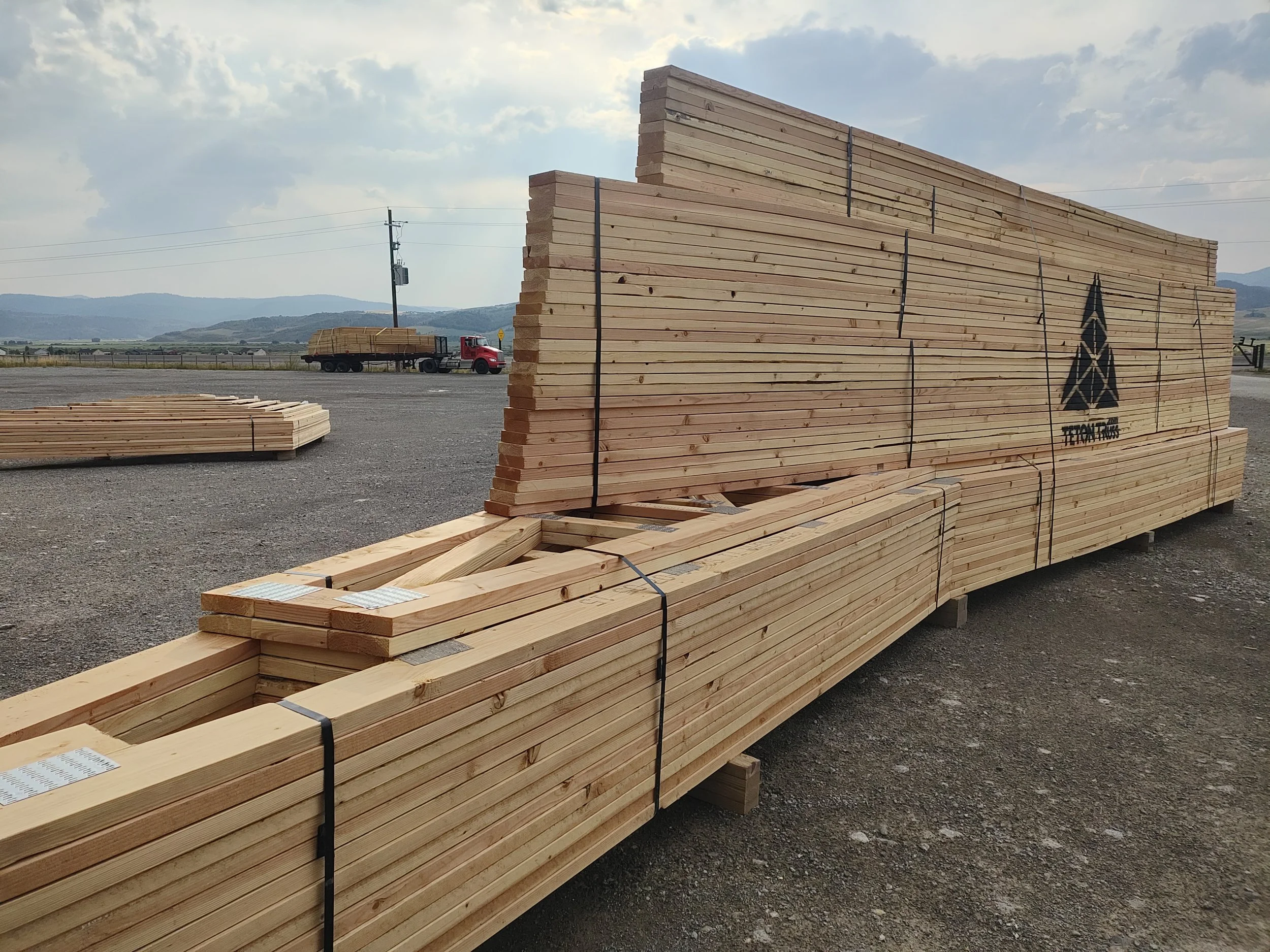 Stacks of lumber being stored outdoors on a gravel lot with a truck and hills in the background, under a partly cloudy sky.