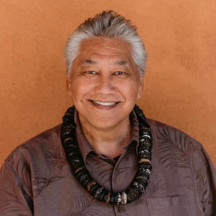 Smiling man with grey hair wearing a dark necklace and patterned shirt against orange background.