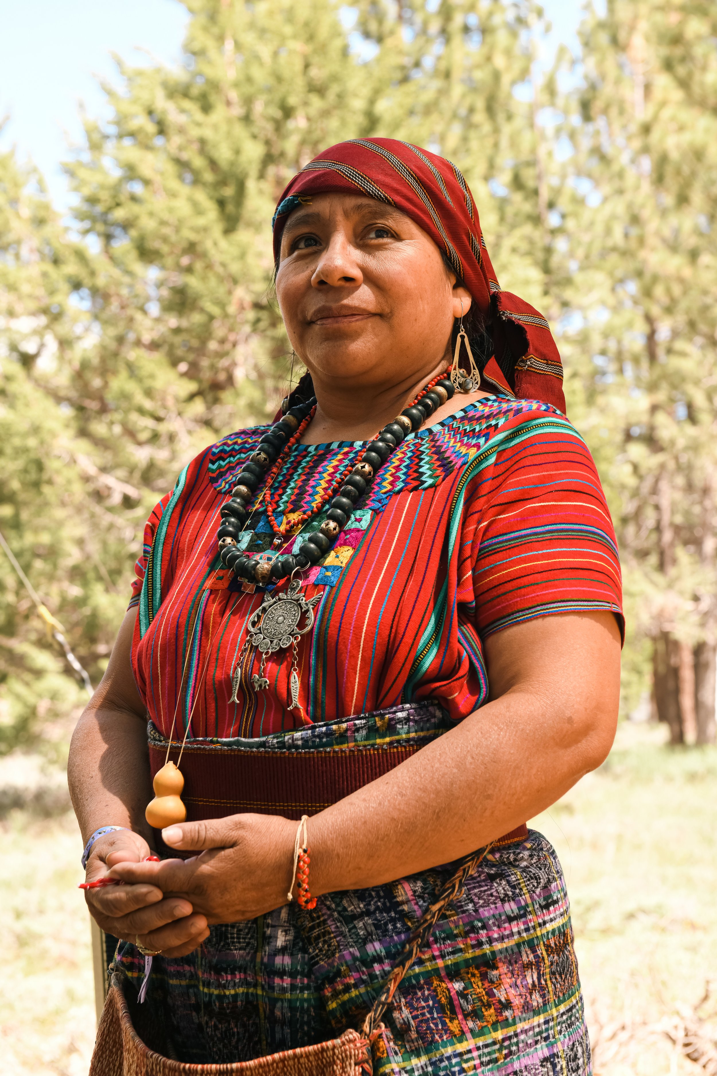 Indigenous woman in traditional clothing with colorful patterns, wearing jewelry and a headscarf, standing outdoors with trees in the background.
