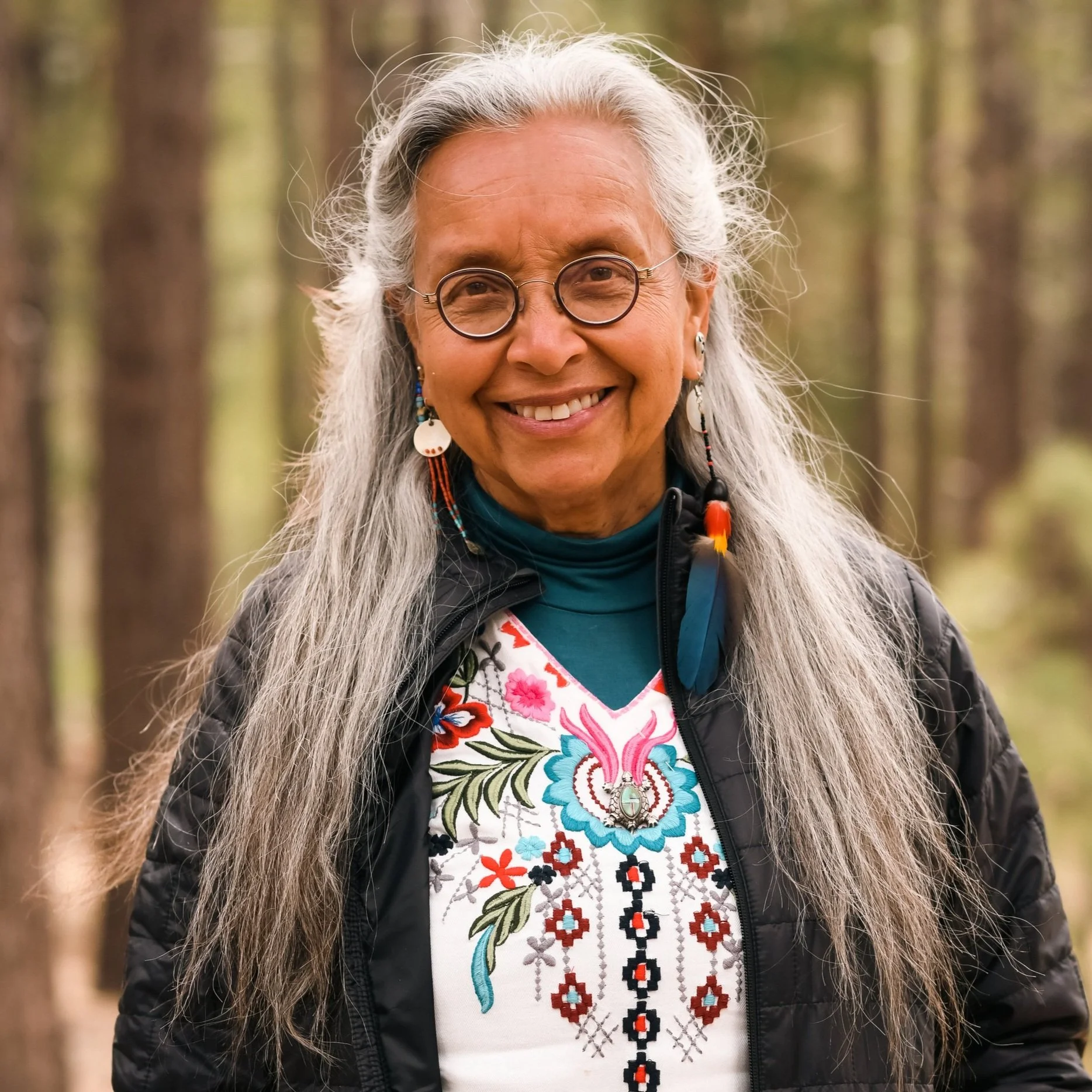 Smiling woman with long gray hair, wearing glasses, earrings, a black jacket, and a floral-patterned shirt, standing in a forest.