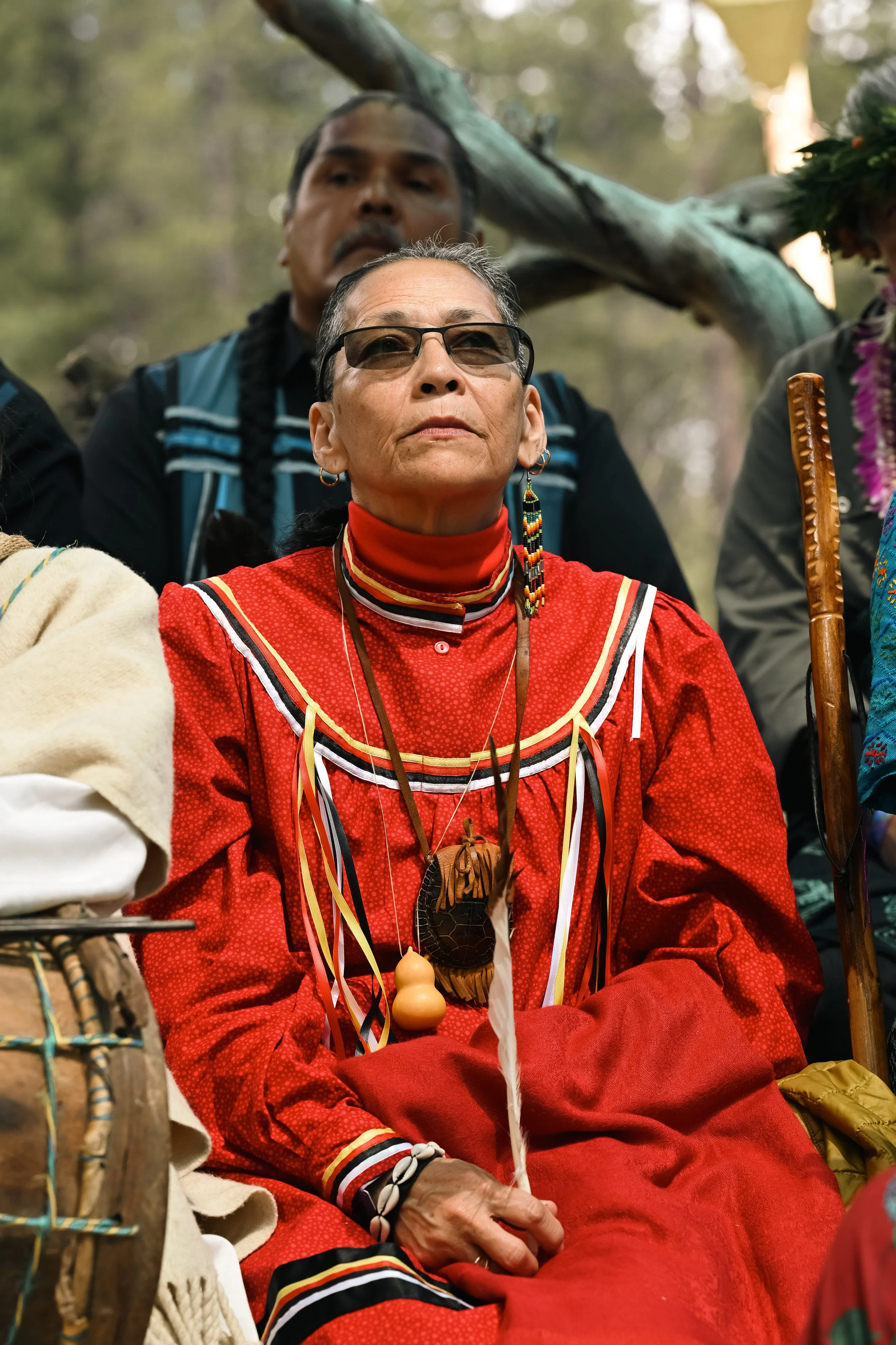Person wearing traditional red attire with colorful accessories and holding a small gourd, seated outdoors.