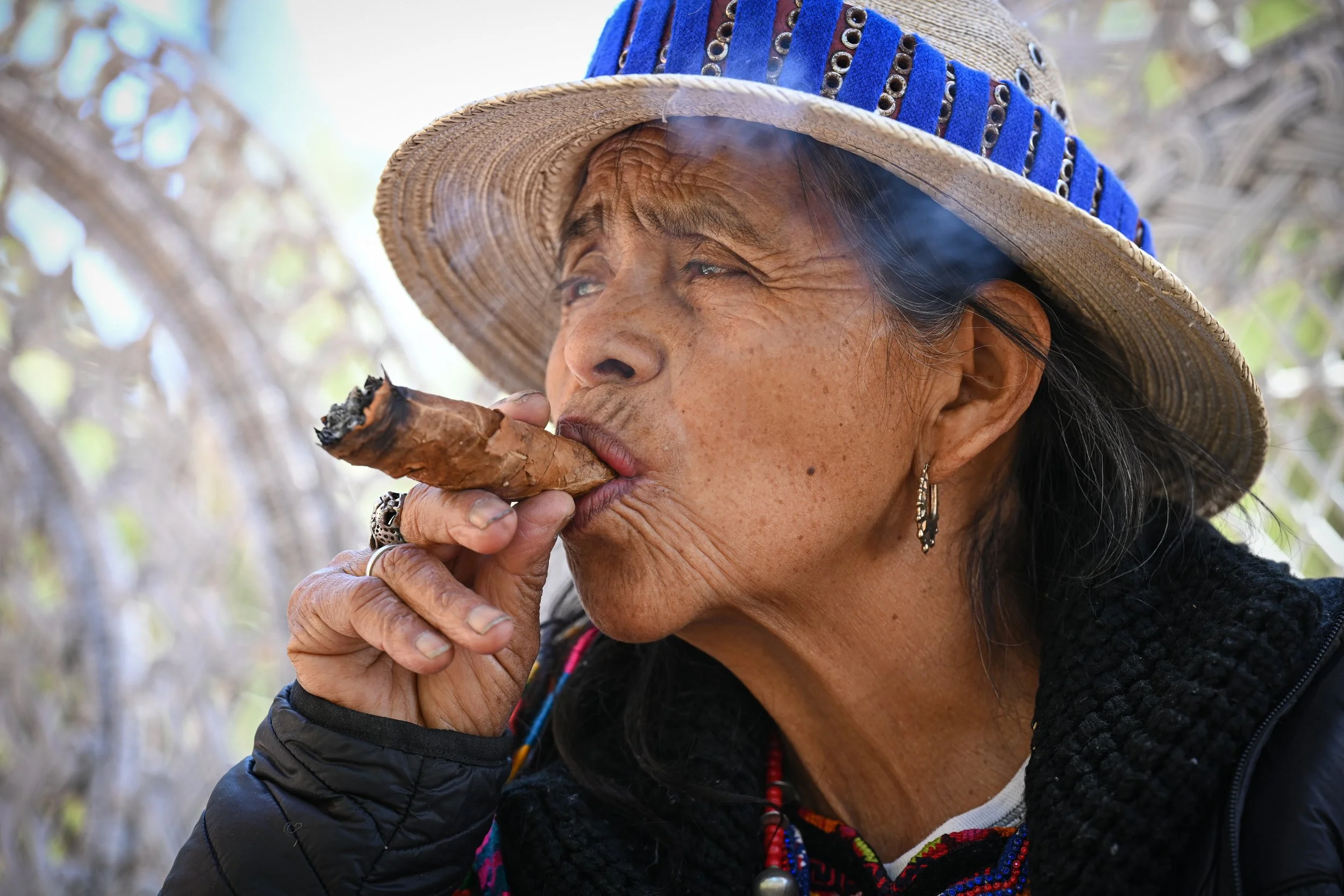 Elderly woman smoking a cigar, wearing a straw hat with blue accents and earrings.